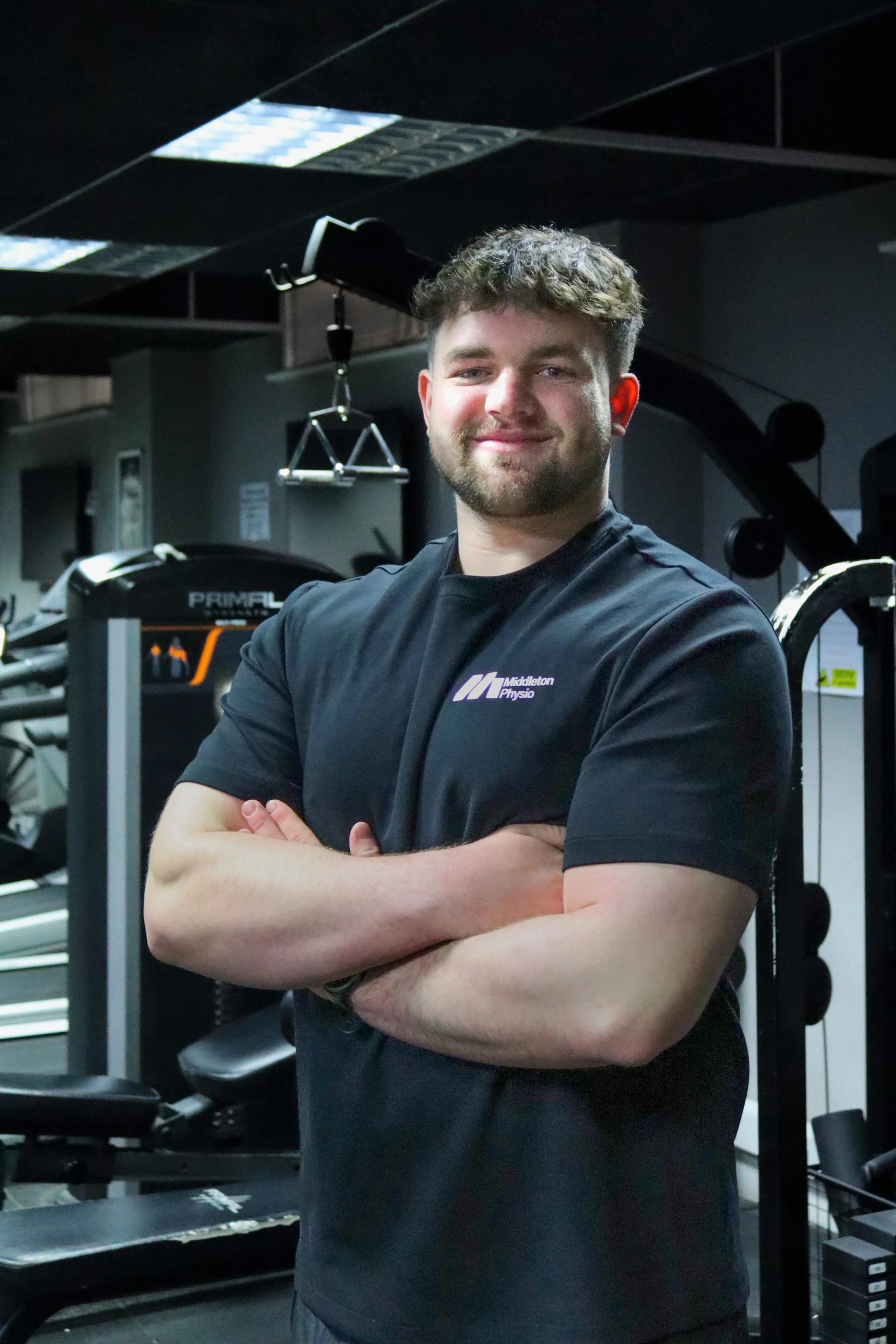 A young man with short curly hair and a beard standing in a gym, smiling with arms crossed, wearing a black shirt with a logo that says 'Middleton Physio.' Gym equipment like exercise machines and weights are visible in the background.
