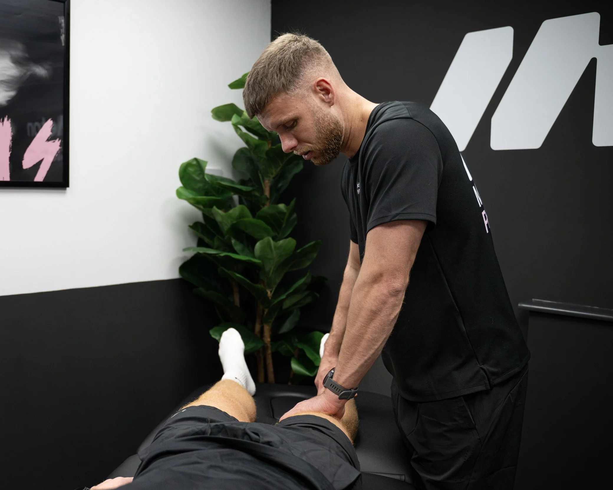 A physical therapist or trainer performs a treatment or exercise on a male patient lying on a black massage table in a modern, professional clinic with black and white walls and a large green plant.