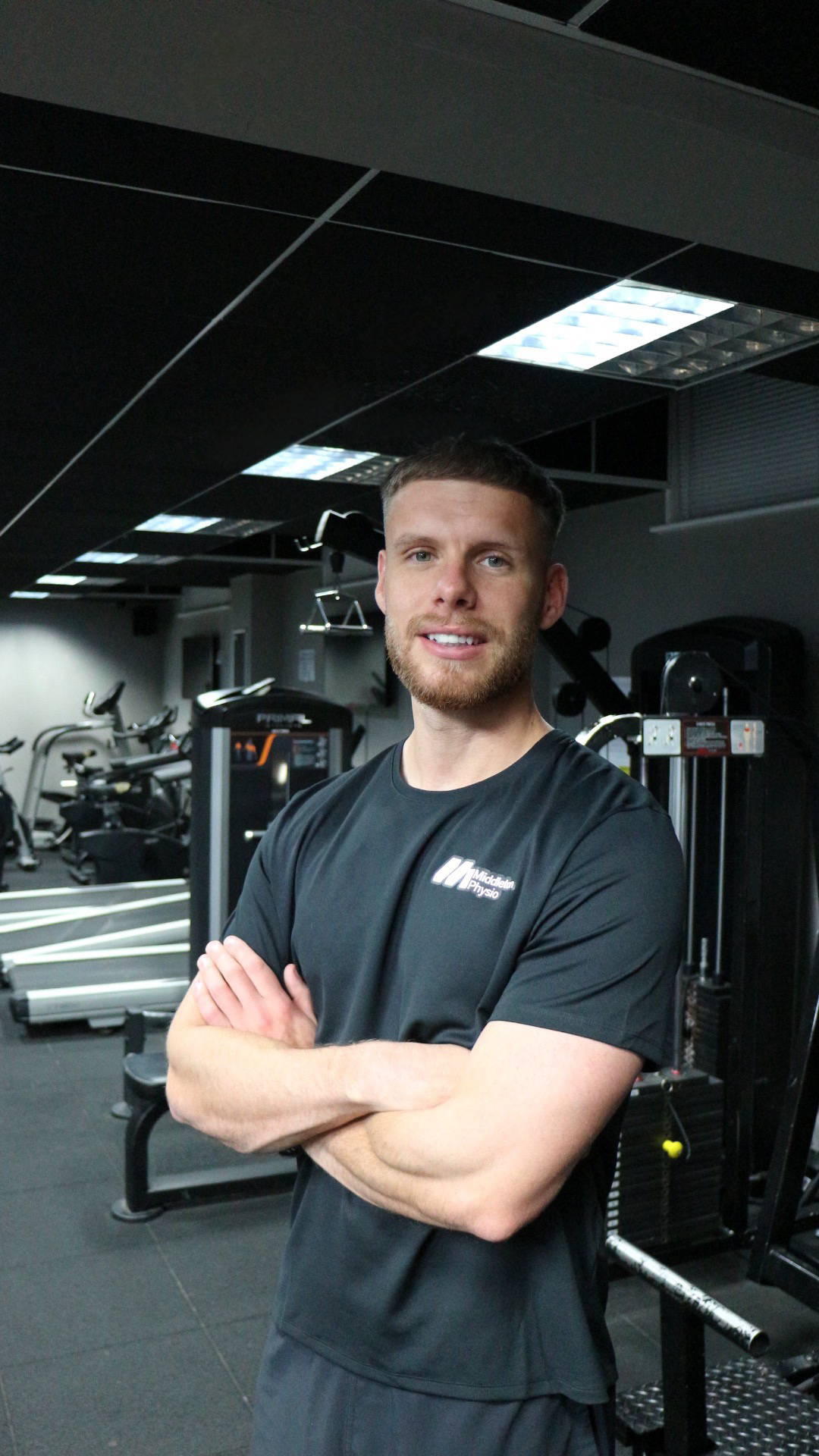 A man with short hair and a beard, wearing a black t-shirt with 'Middleton Physio' on it, standing with arms crossed in a gym filled with workout equipment and exercise machines.