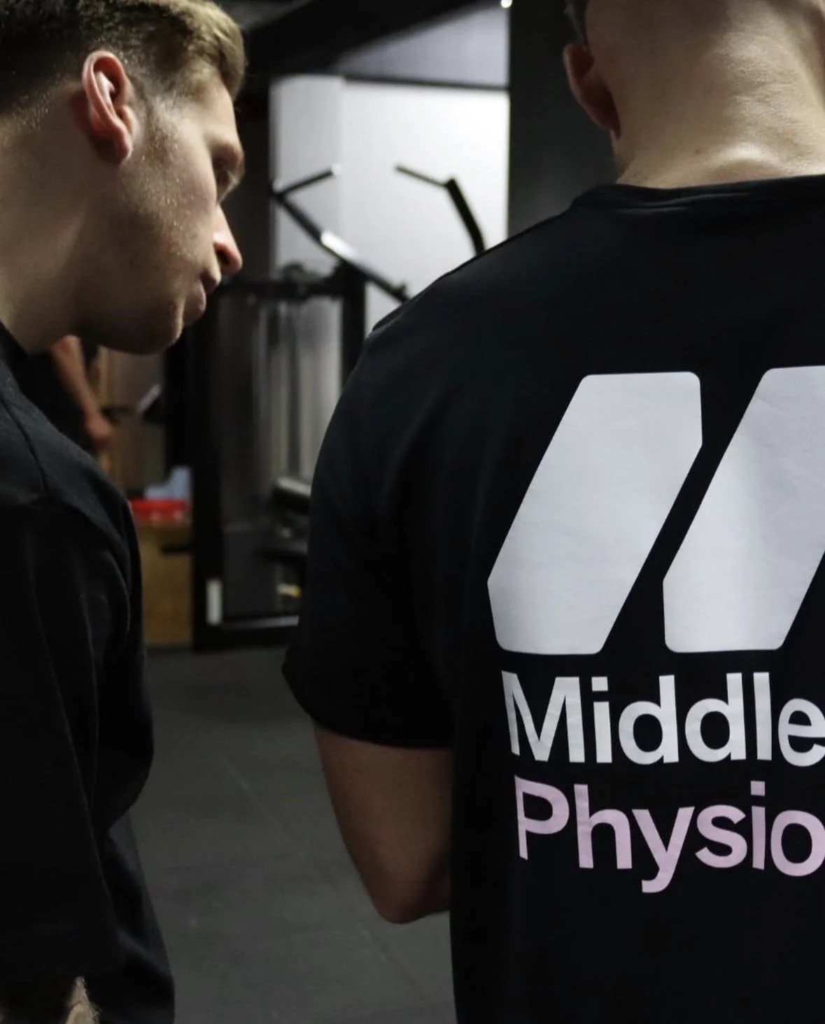 Two people standing close together in a gym, one wearing a black T-shirt with 'Middle Physio' printed on the back.