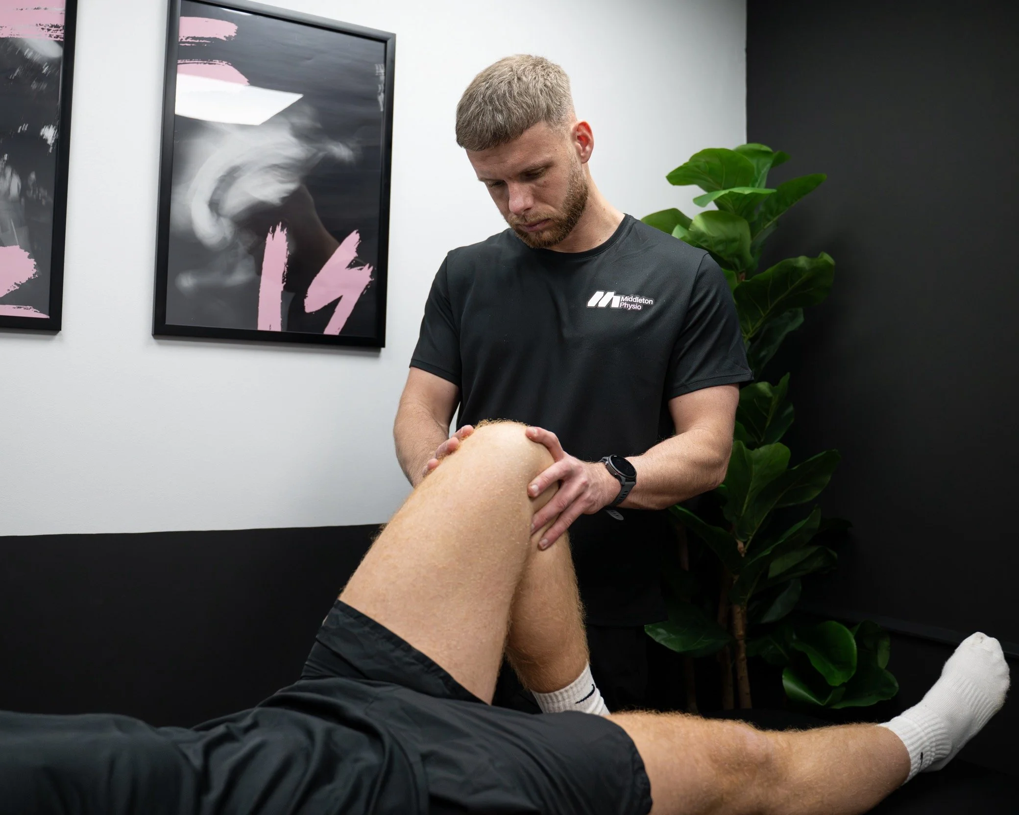 A physical therapist examines a patient's knee in a clinic. The therapist wears a black shirt, and the patient is lying down with one leg bent. There is a plant and framed artwork on the wall behind them.