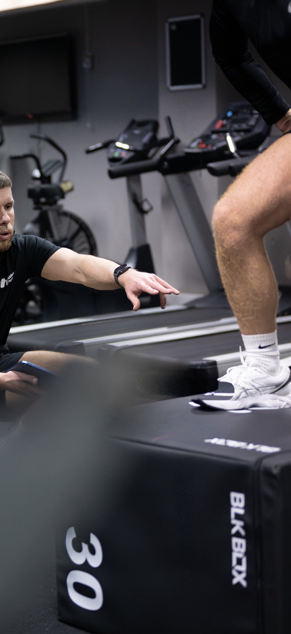 A trainer assists a person running on a treadmill during a workout session at a gym, with exercise equipment in the background.