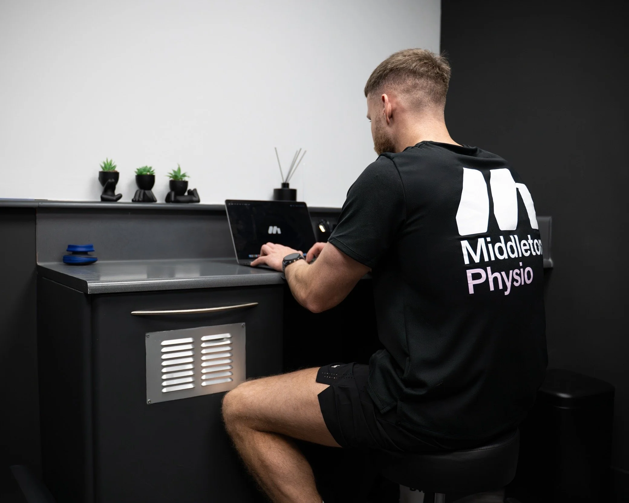 A young man with short hair and a beard, wearing a black Middleton Physio t-shirt and shorts, sitting on a black round stool at a black desk, typing on a laptop in a room with black and white walls, small potted plants on the desk, and a black diffuser with reed sticks.
