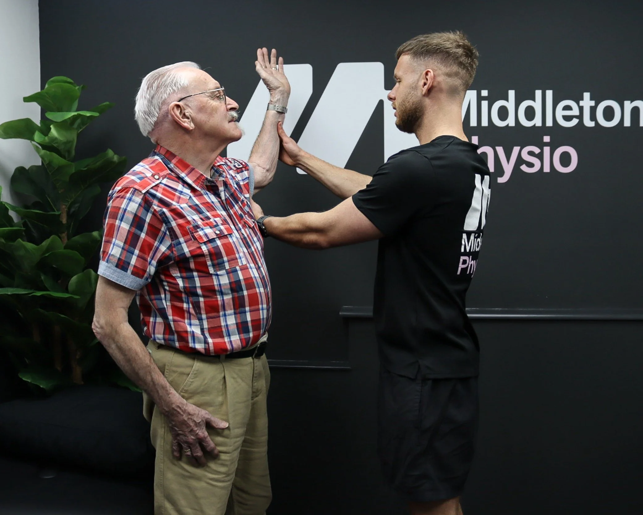 An elderly man and a younger man engaging in a physical therapy or rehabilitation session at Middlesex Physio, with the younger man supporting the elderly man's arm while they face each other, both appearing focused.