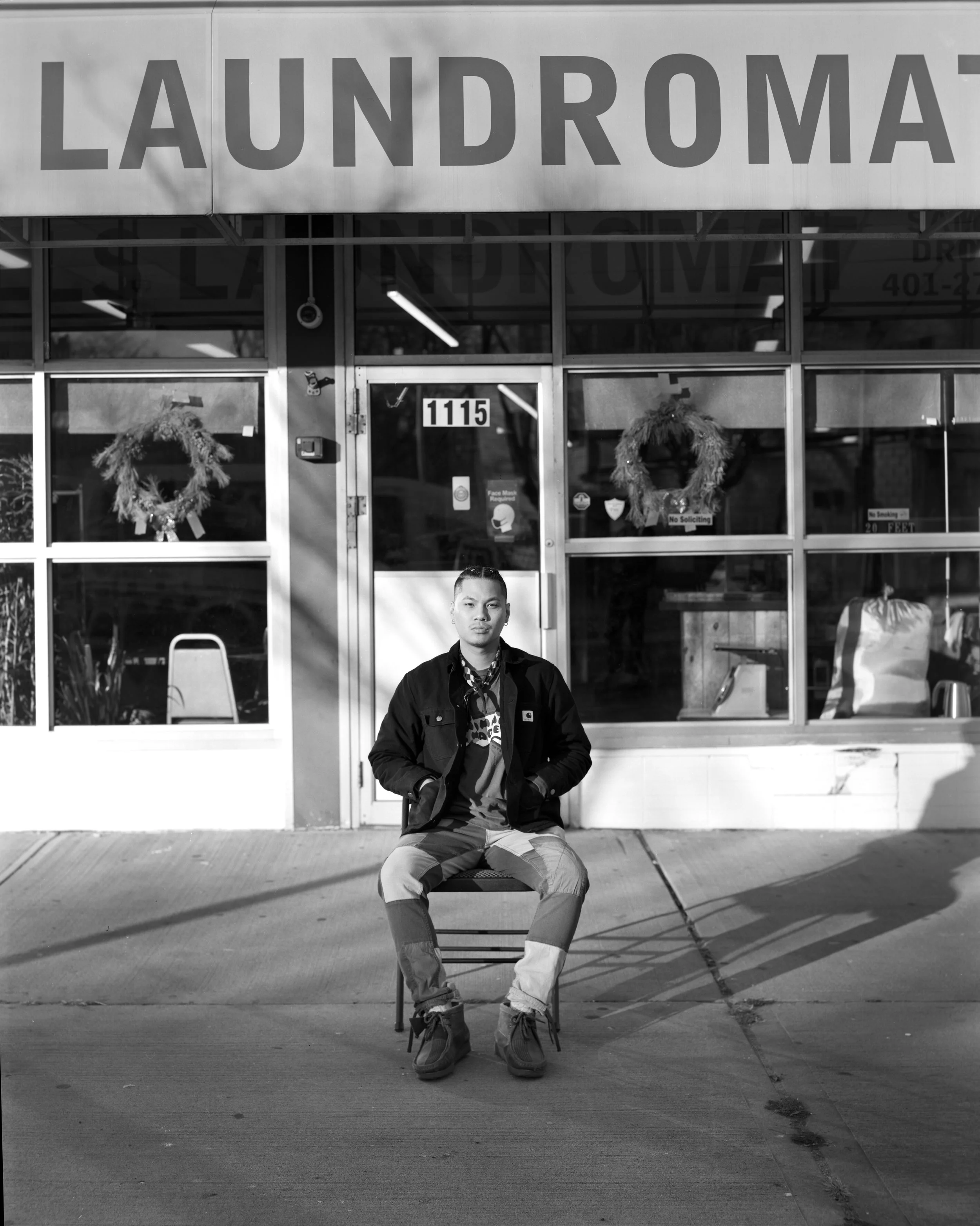 A young man sitting on a chair outside a laundromat with large windows and festive wreath decorations on the door.