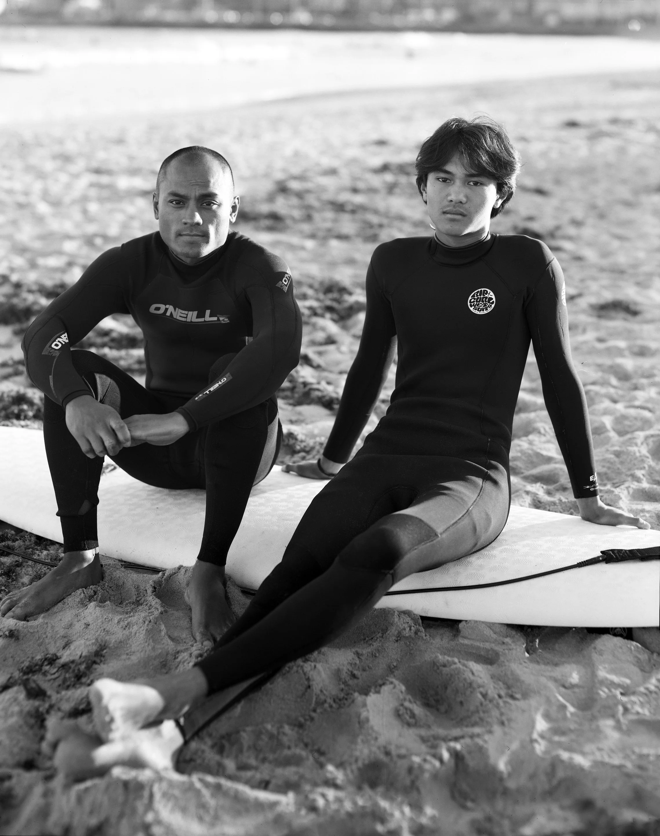 Two young men wearing wetsuits on a beach, one sitting with his elbow on his knee and the other relaxed on a surfboard, both looking at the camera.