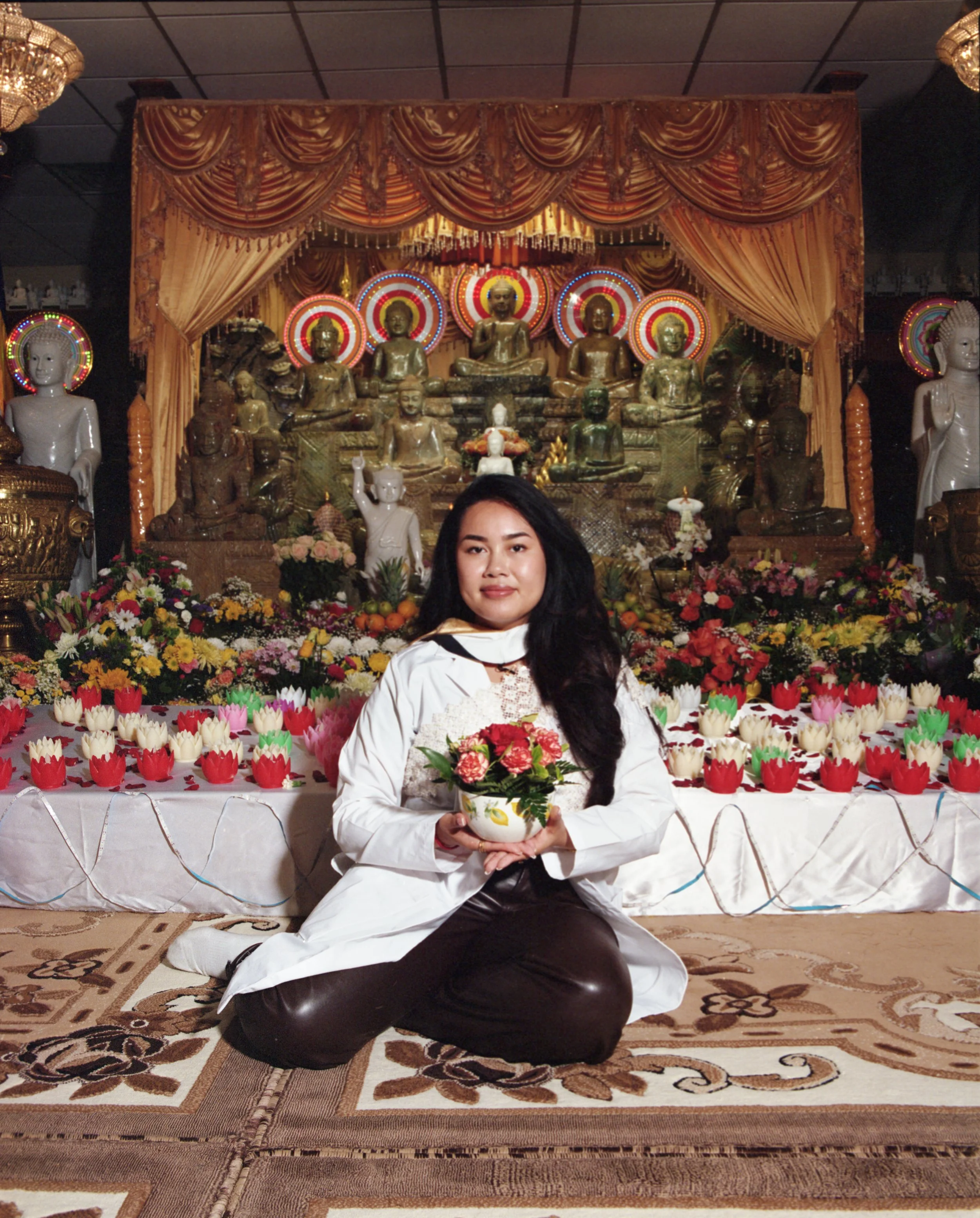 A woman in traditional attire holding a flower arrangement, kneeling in front of a Buddha altar decorated with flowers and statues.