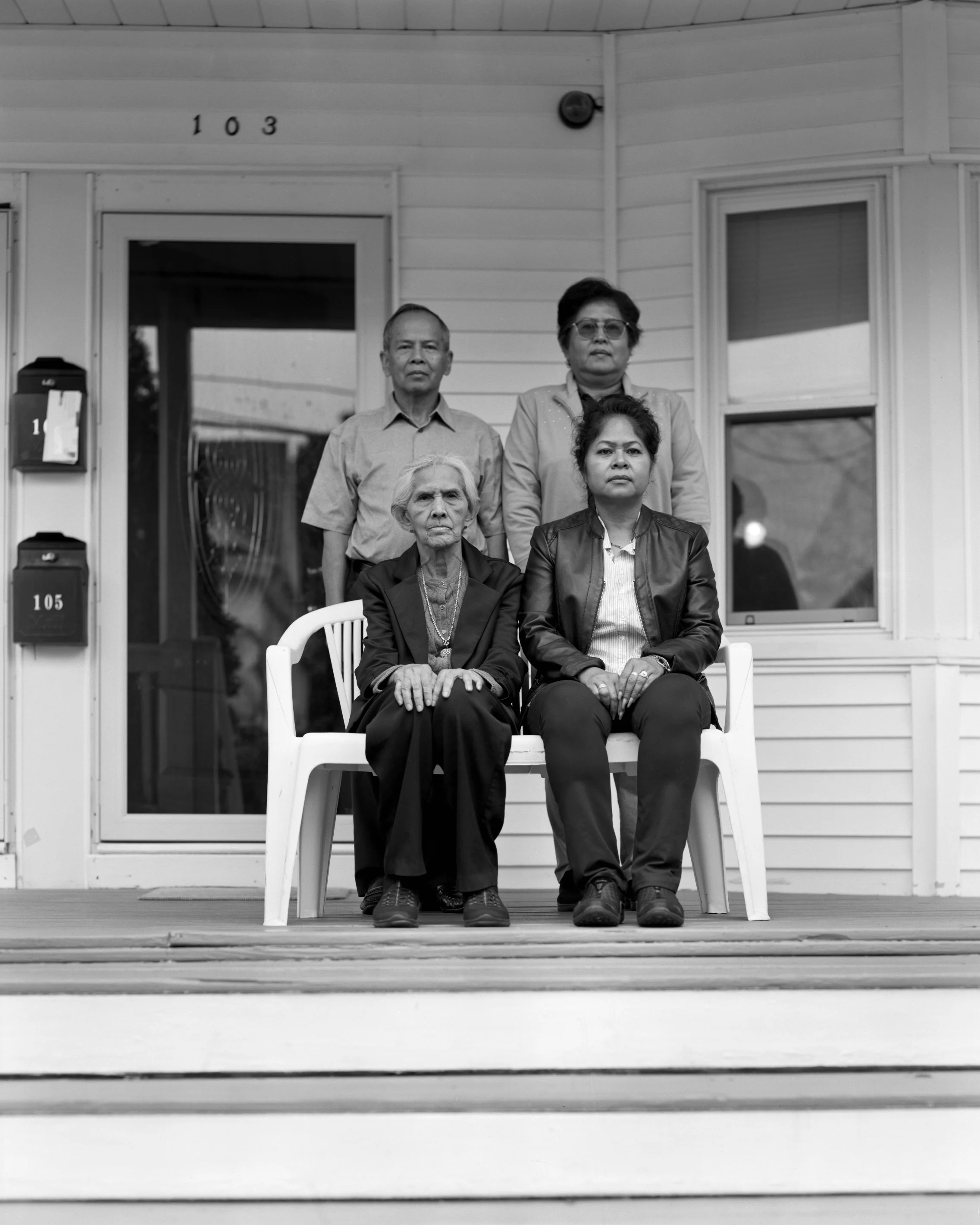 A black and white photo of five people on a porch in front of a house. Two women are seated in the front, one elderly and one middle-aged, and three men and women stand behind them. The house behind them has the number 103 and 105 on the wall next to