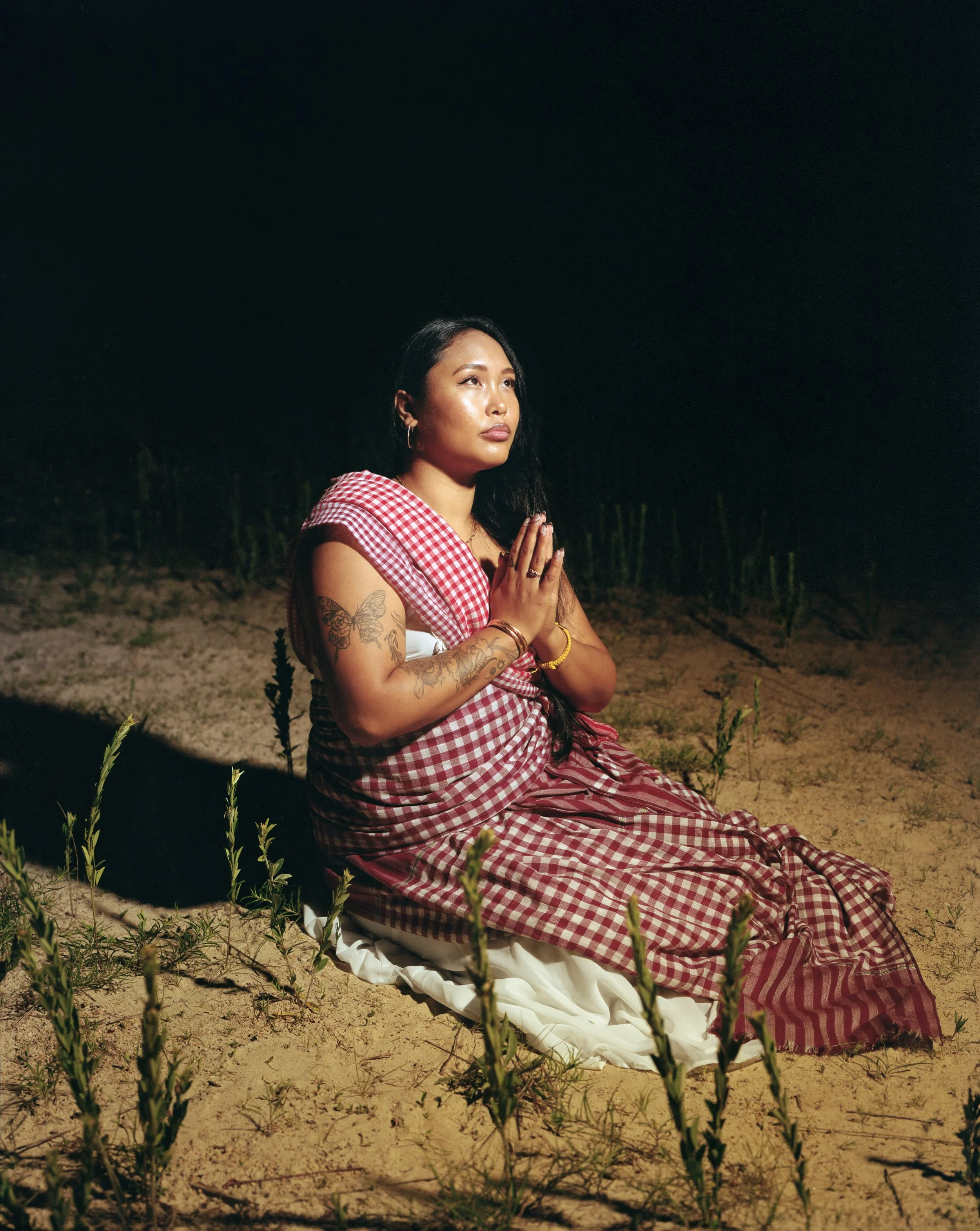A woman praying outdoors at night, kneeling on the sandy ground with her hands pressed together and eyes closed.