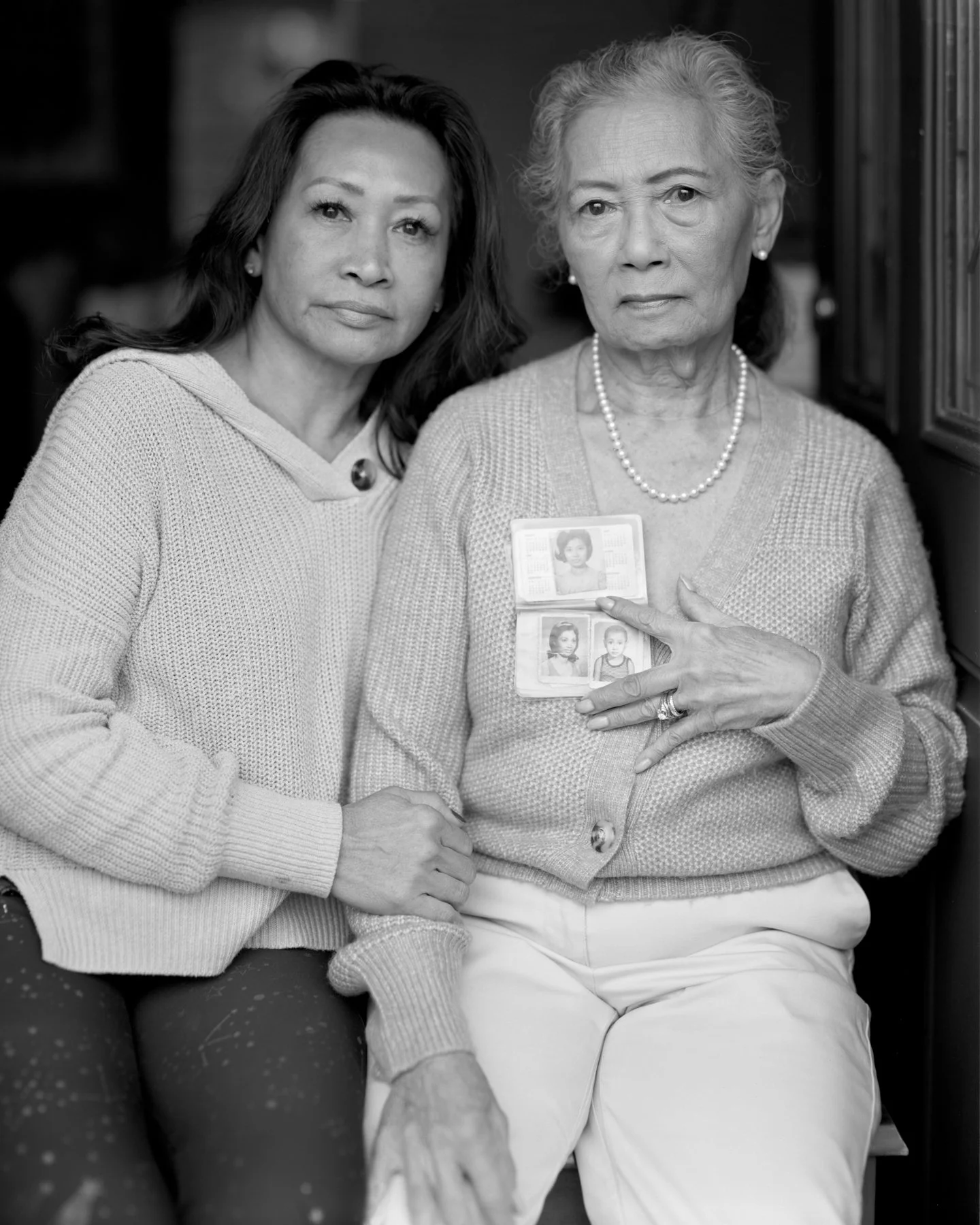 A younger woman and an elderly woman sitting close together, the elderly woman pointing to her identification badge with photos of children.