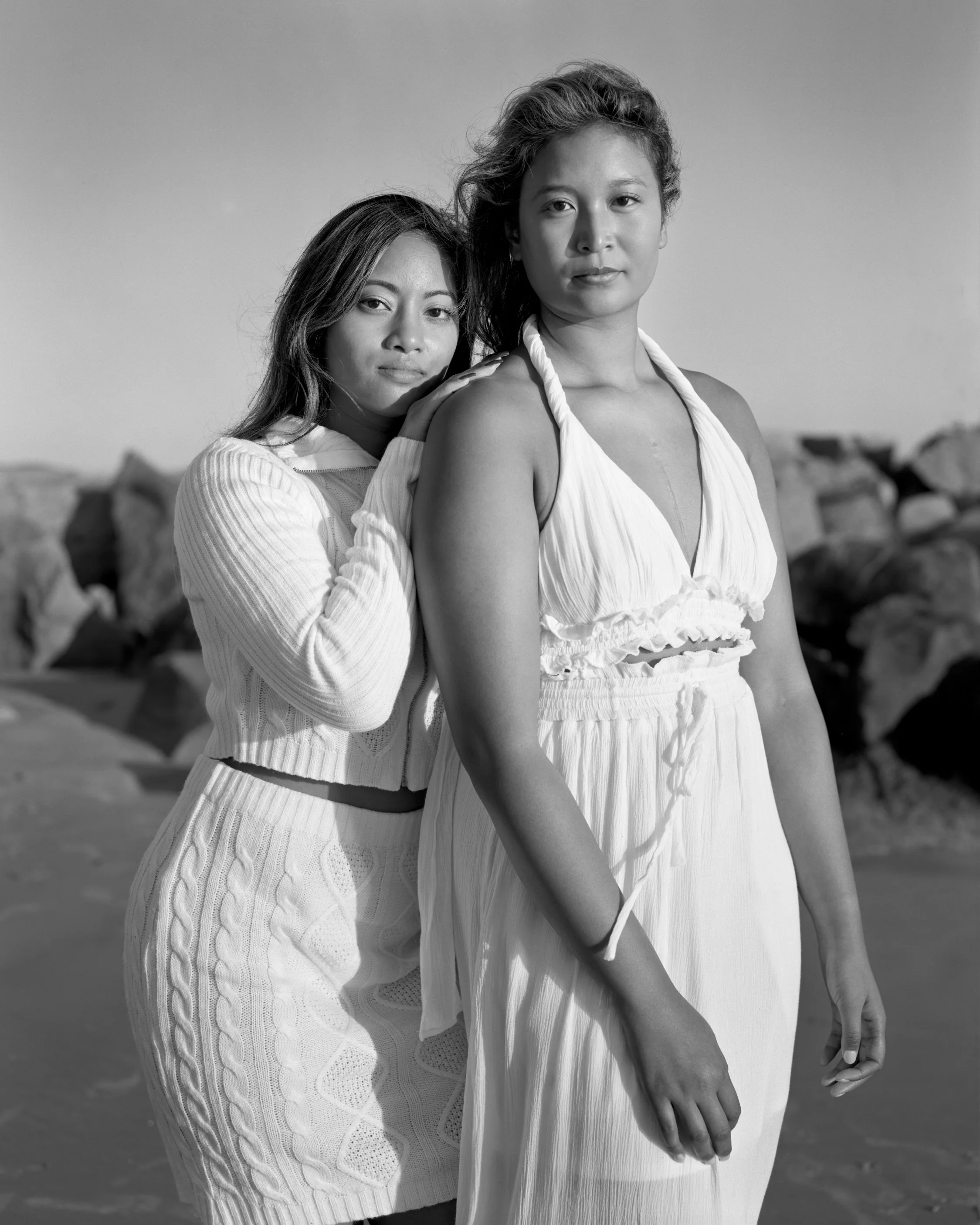 Two women standing on a rocky beach, one resting her hand on the other's shoulder. Both are dressed in light-colored, summery clothing and looking at the camera.