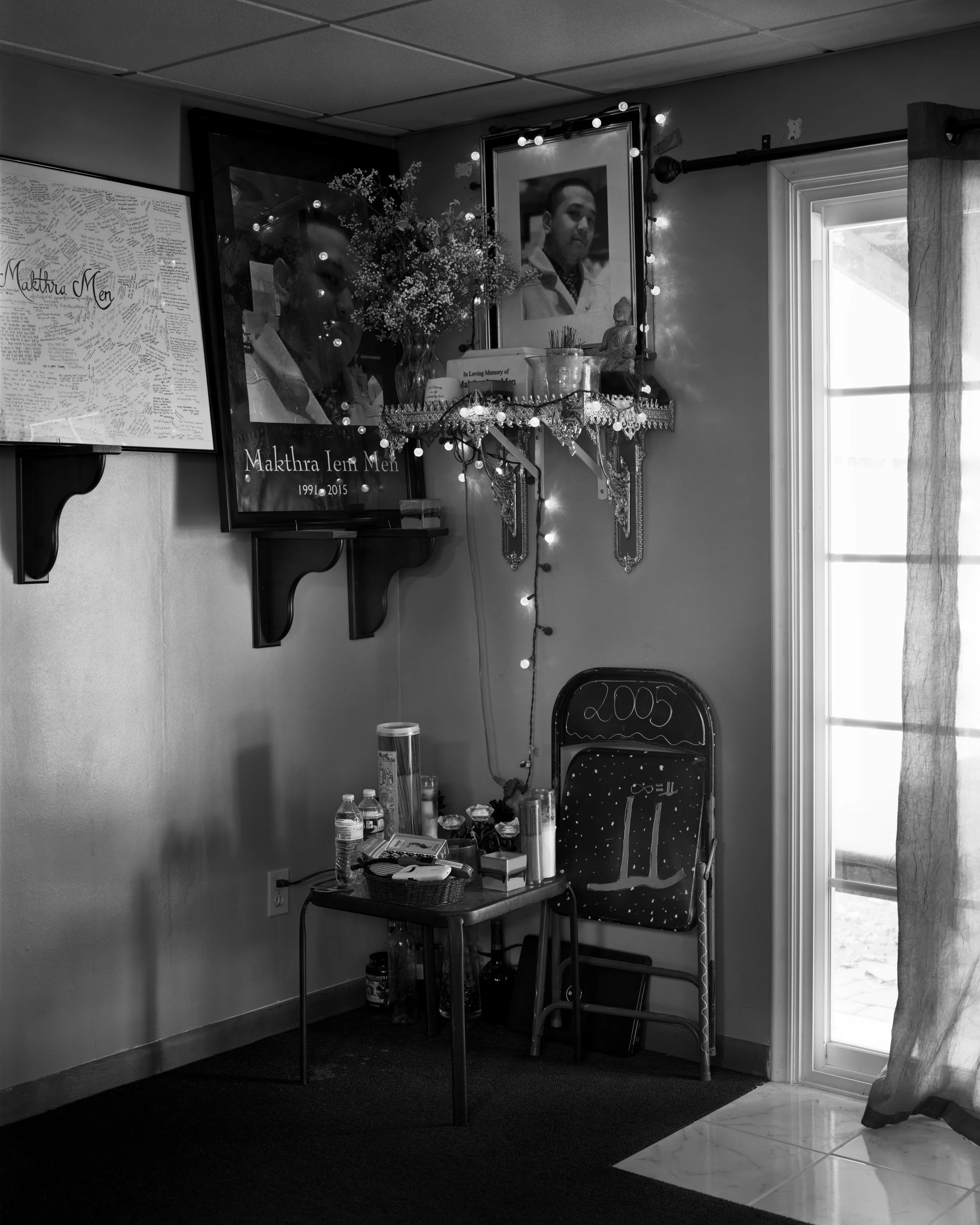 A memorial corner with framed photographs, a small table with water bottles and candles, a folding chair with "2005" written on it, and decorative lights on the wall.
