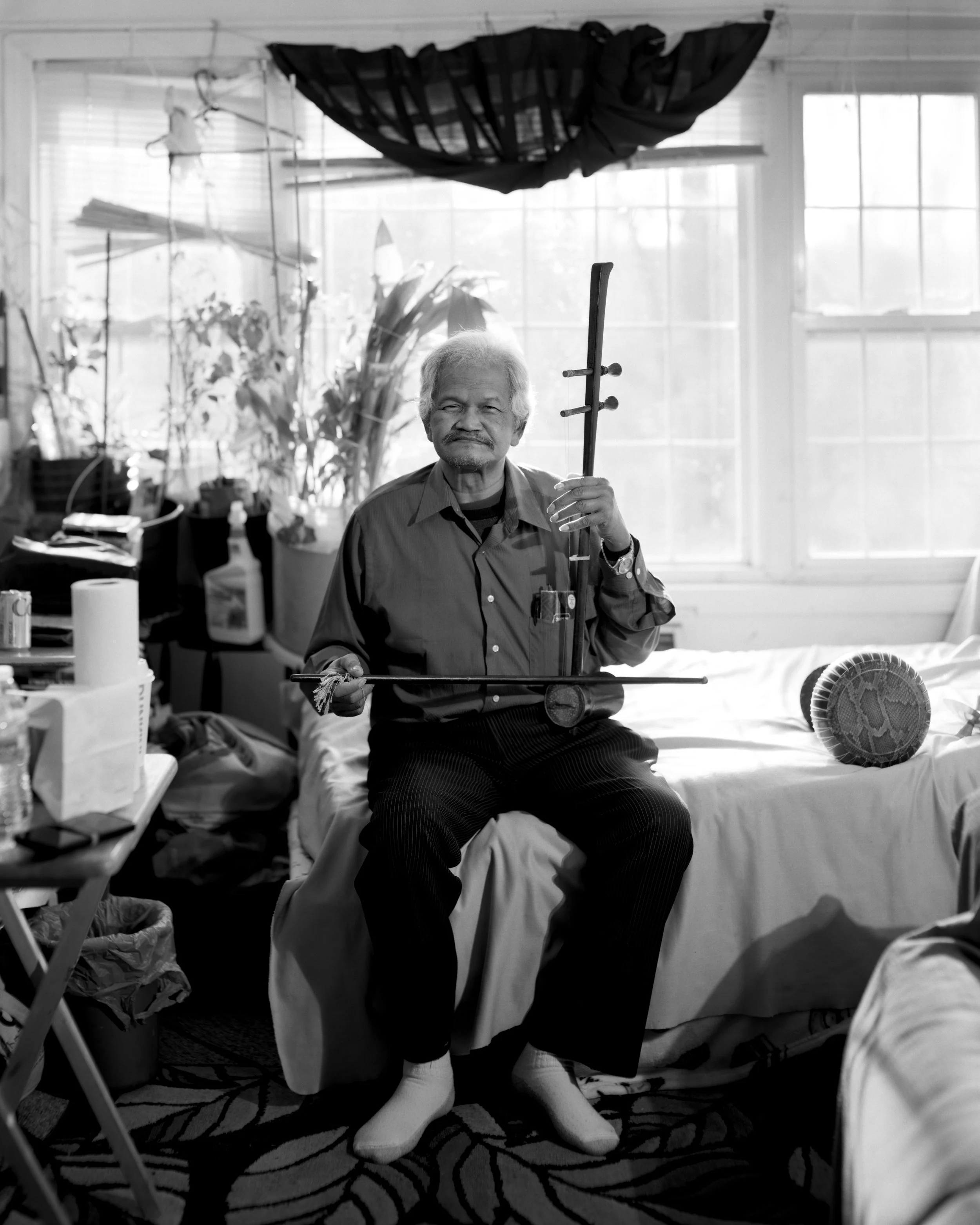 An elderly man with gray hair and a beard, sitting on a bed in a cluttered room, holding a traditional musical instrument. The room has large windows with blinds, various plants, and personal items around.
