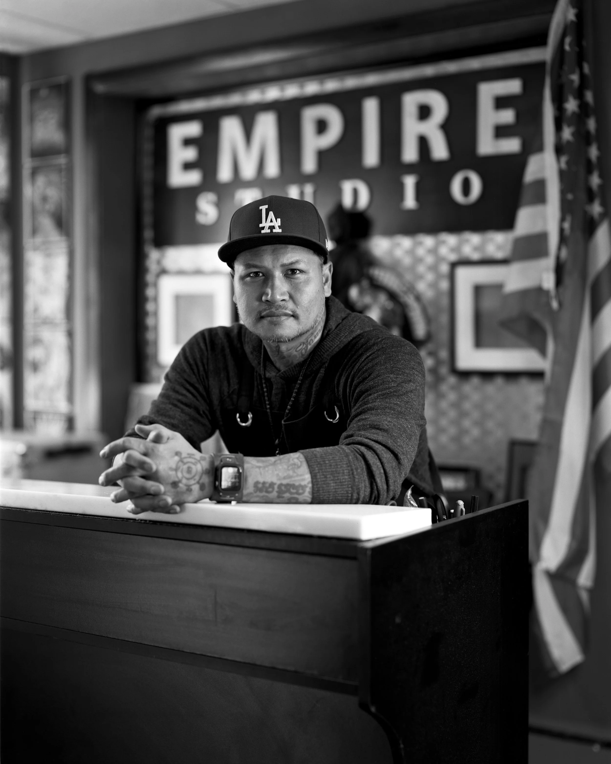 A man with tattoos and a cap sitting at a reception desk in a studio, with the sign 'EMPIRE STUDIO' and an American flag in the background.