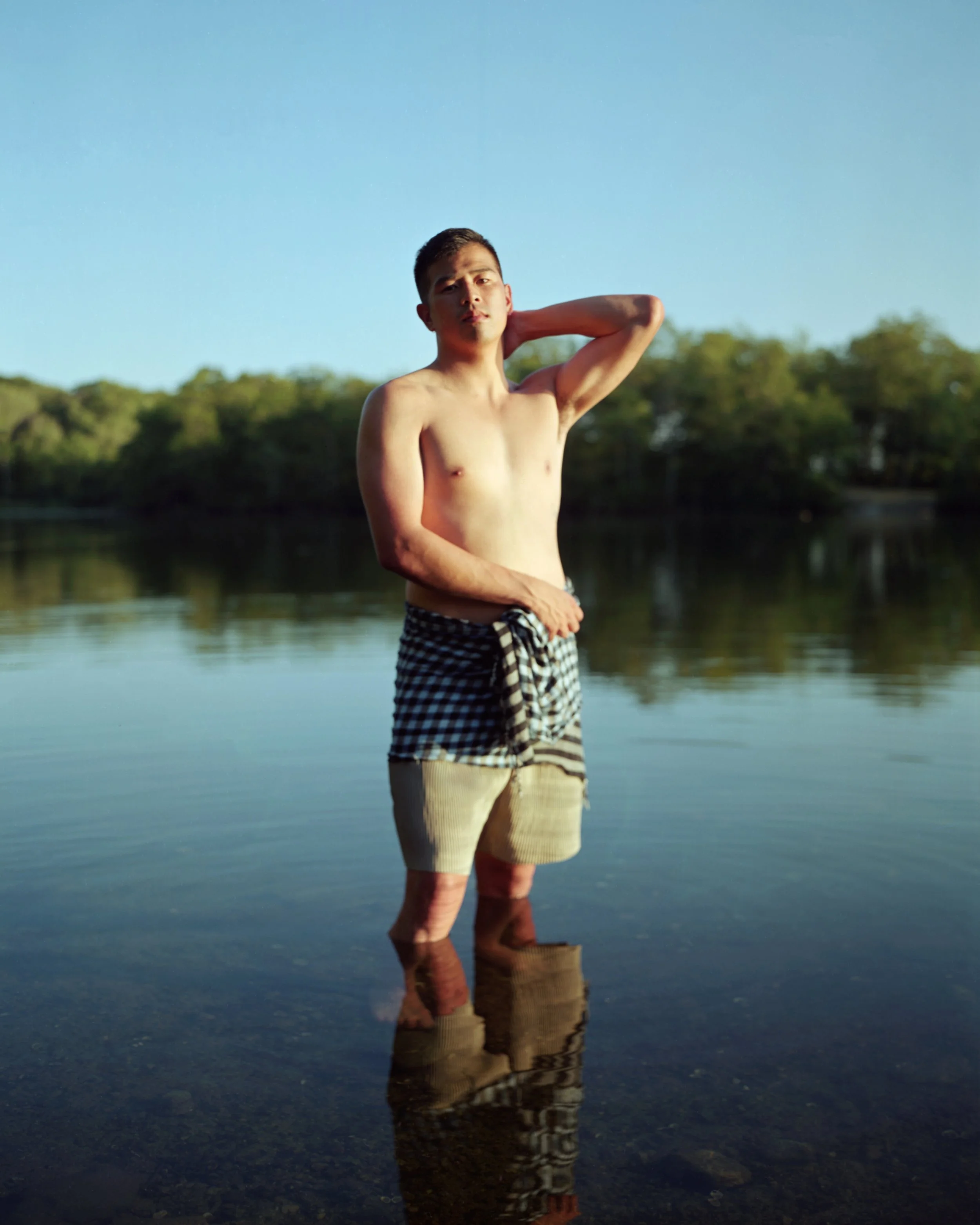 A shirtless man stands in shallow water at a lake, with trees and a clear blue sky in the background, posing with one hand behind his head.