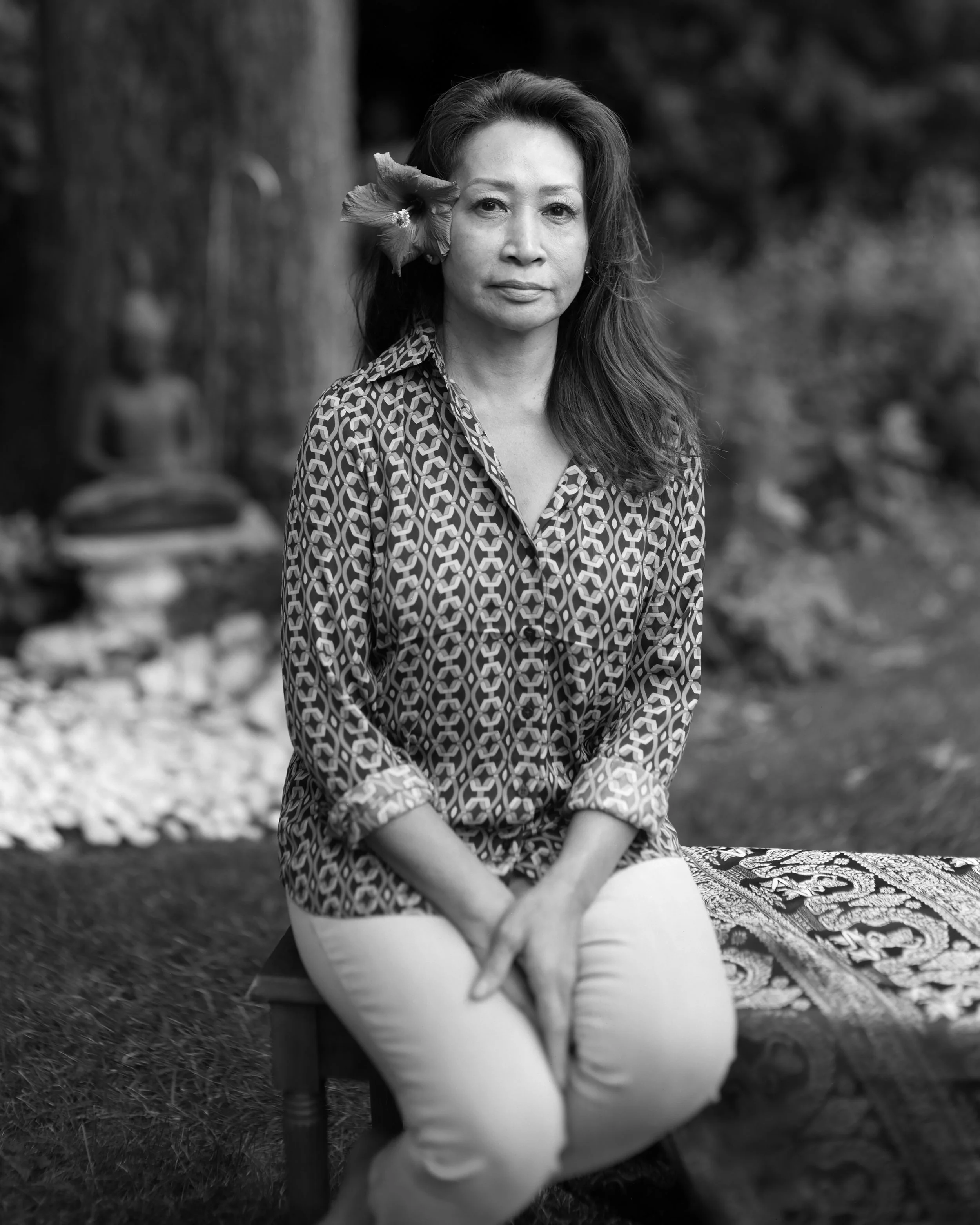 A woman sitting outdoors on a wooden stool with her hands in her lap, wearing a patterned shirt, with a flower in her hair. The photo is in black and white with a blurred natural background.