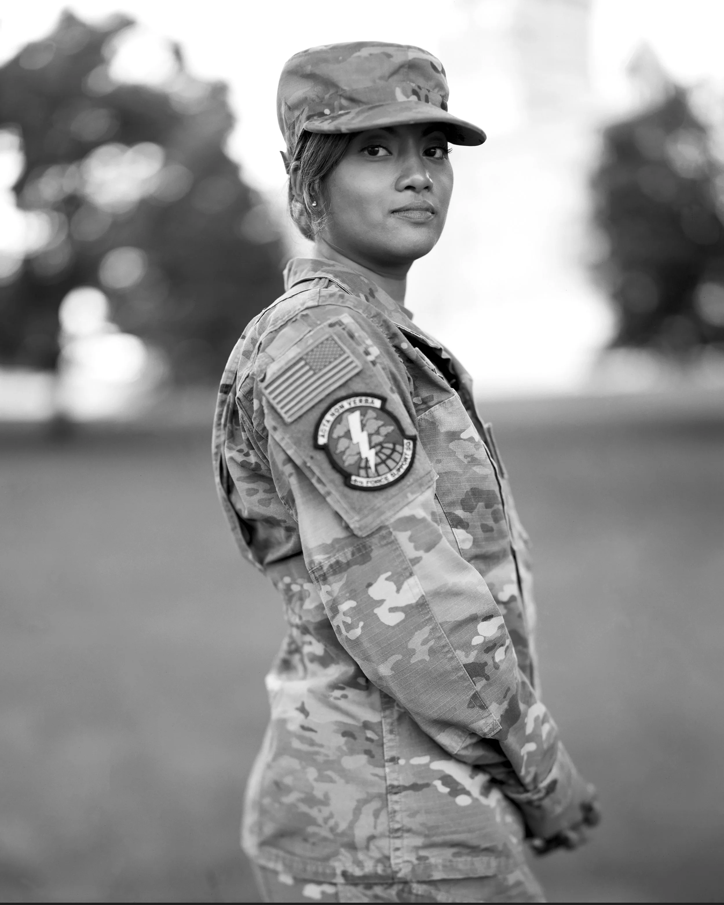 Black and white photo of a woman in military camouflage uniform and cap, standing outdoors with blurred trees in the background, looking at the camera.