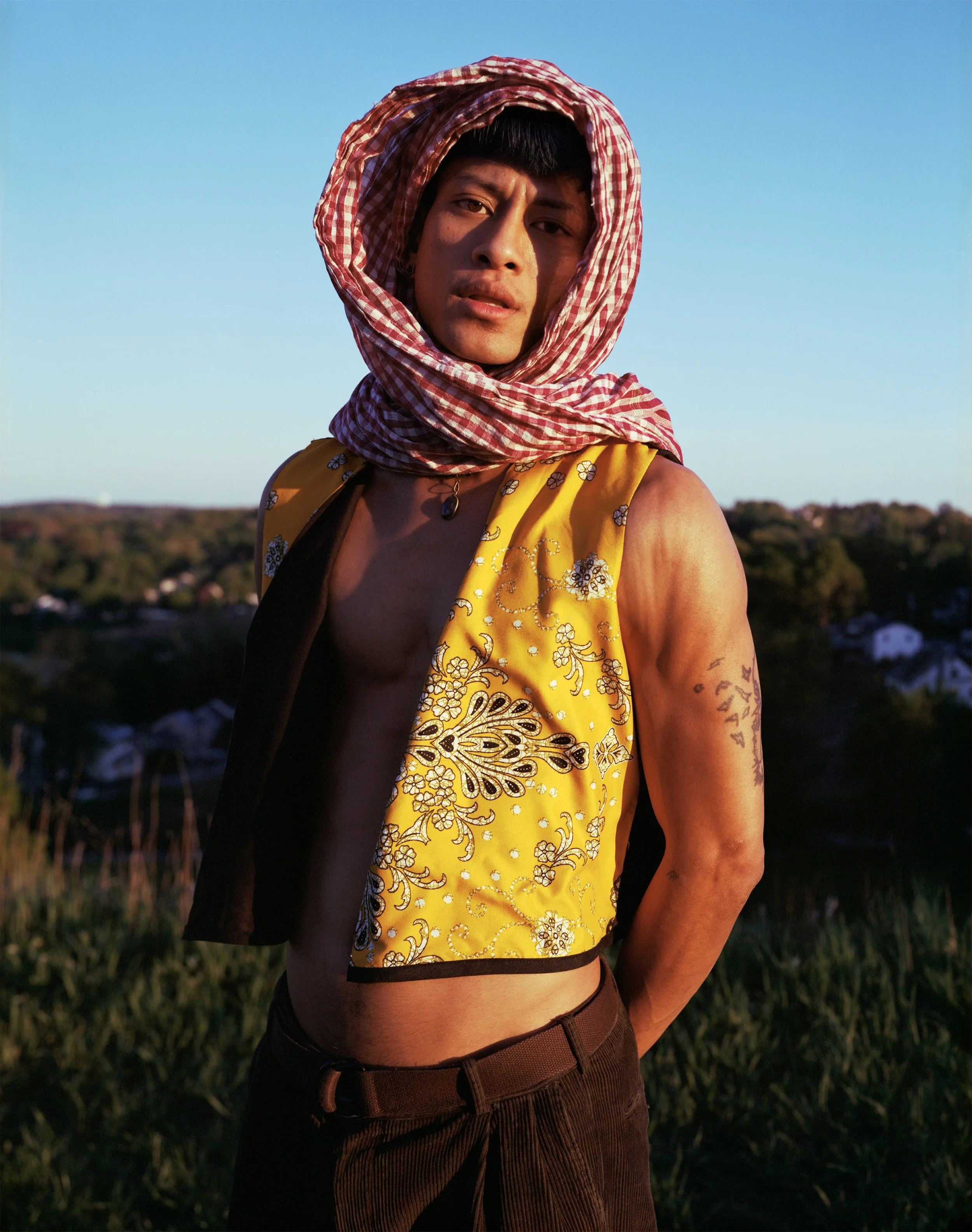 A young man with a scarf around his head, wearing a sleeveless yellow and black vest, standing outdoors during sunset with a landscape of trees and houses in the background.