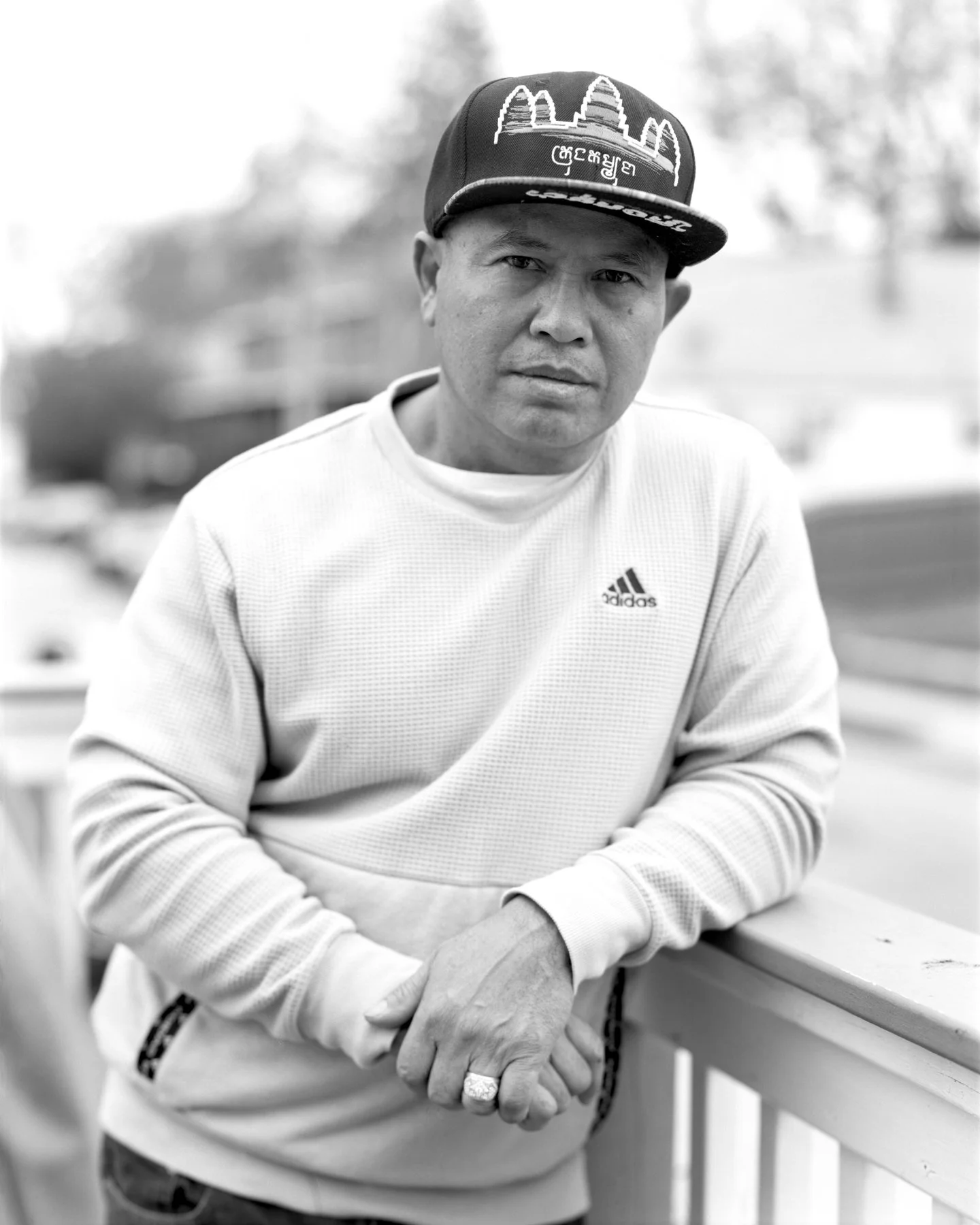 A man wearing a flat-bill cap and a white Adidas sweatshirt, leaning against a railing outdoors, with a slightly serious expression.