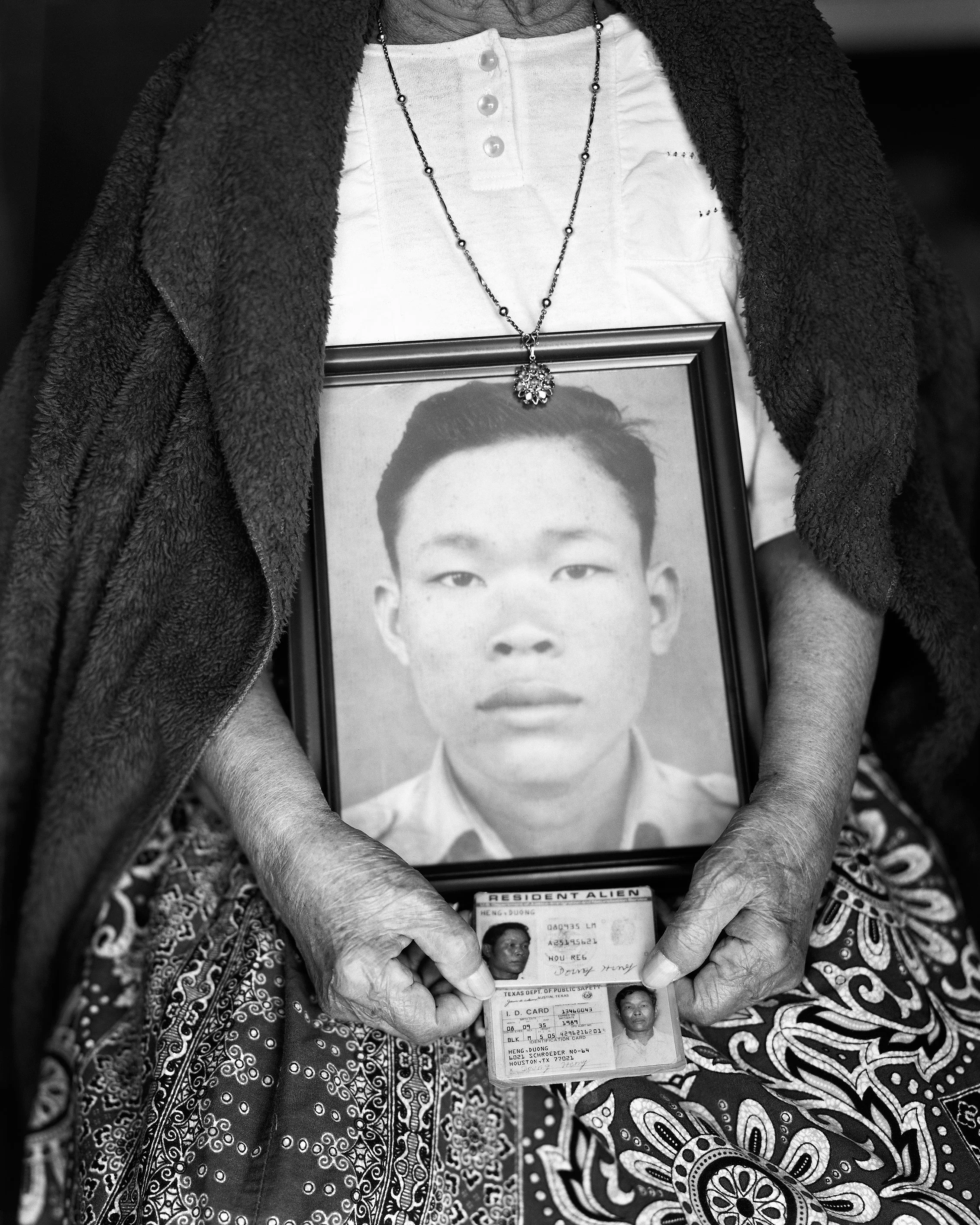 An elderly woman holding a framed black-and-white portrait of a young man and an ID card.