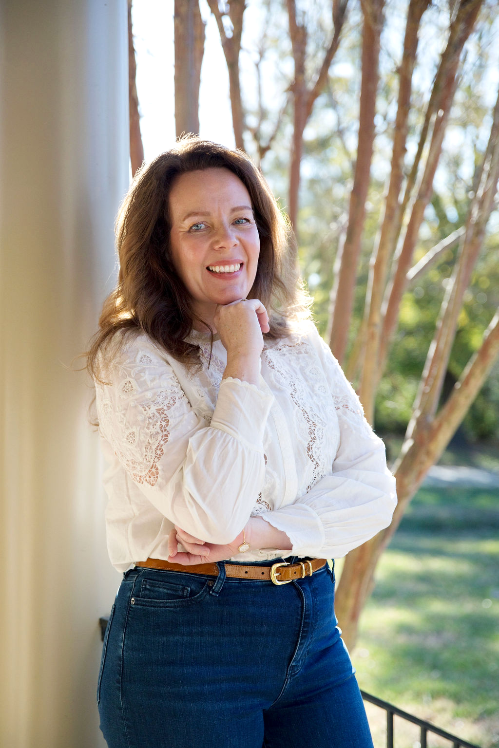 A woman with dark brown hair, wearing a white lace blouse and dark blue jeans, smiling outdoors with trees in the background. Anita McGovern Real Estate
