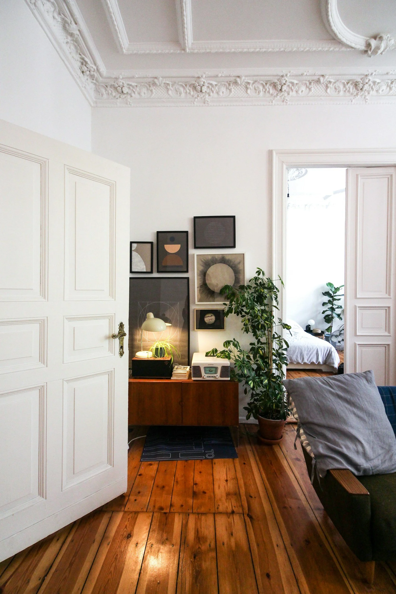 A living room corner with a wooden sideboard, framed artwork, plant, lamp, radio, and doorway leading to a bedroom. Anita McGovern Real Estate