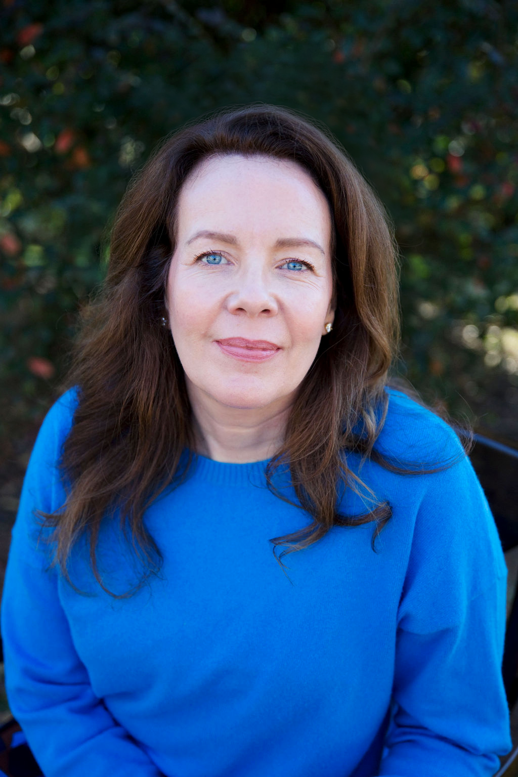 A woman with long brown hair and blue eyes wearing a bright blue sweater, looking directly at the camera outdoors with blurred green foliage in the background. Anita McGovern Real Estate