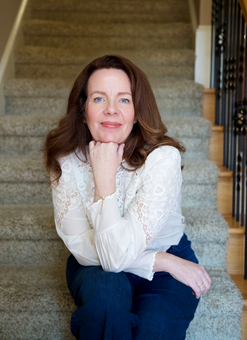A woman with long brown hair, wearing a white lace blouse and dark pants, sitting on a carpeted staircase, resting her chin on her hand and smiling at the camera. Anita McGovern Real Estate