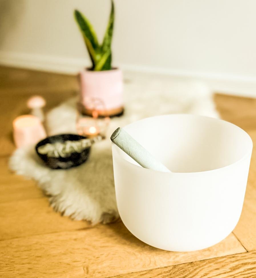A white ceramic bowl with a rolled joint inside it, placed on a wooden table in front of a blurred background with a pink potted plant and ceramic containers.