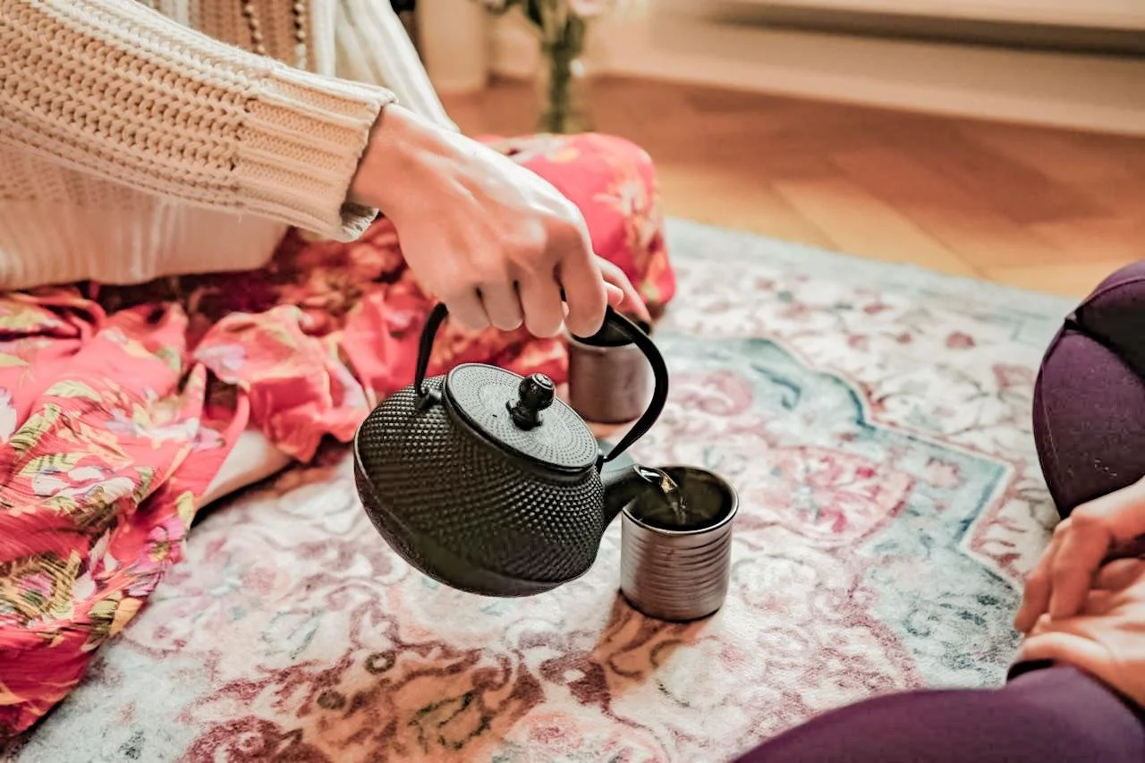 Person pouring hot tea from a black teapot into a metal mug on a patterned rug.