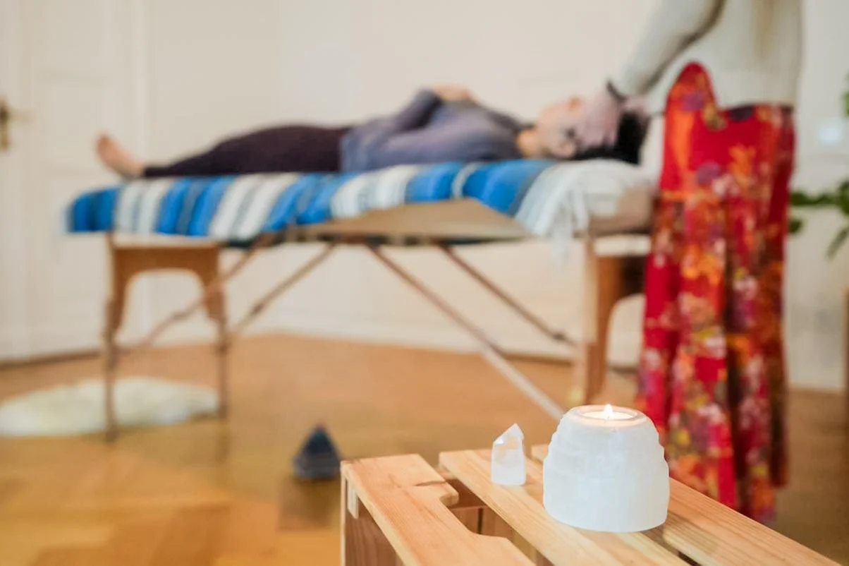 A person lying on an examination table with a cloth over their eyes, with another person holding their head, in a room with a candle and salt lamp on a wooden stand in the foreground.