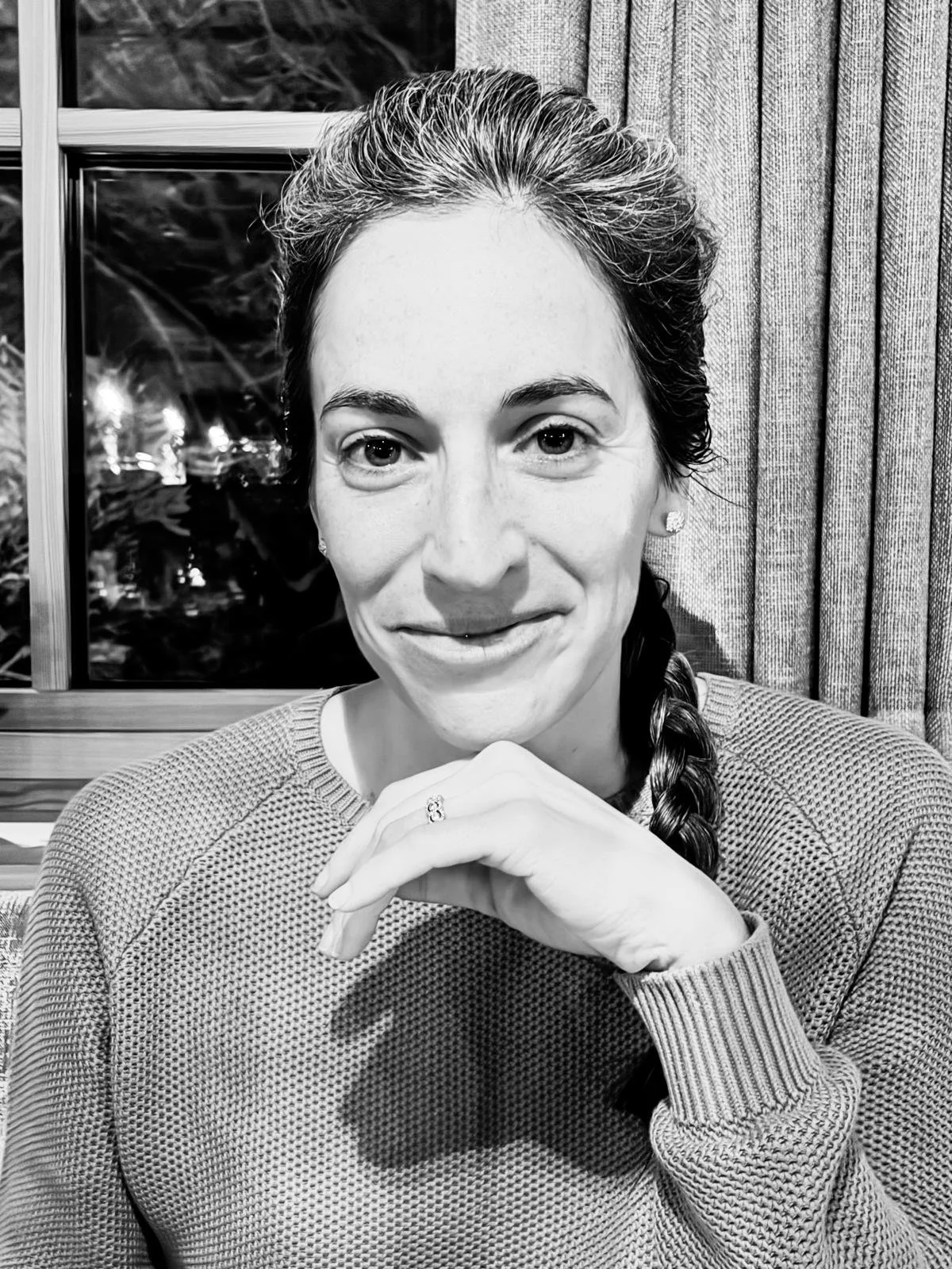 A woman with long braided hair wearing a textured sweater, sitting indoors near a window with curtains, resting her chin on her hand, and smiling slightly at the camera.