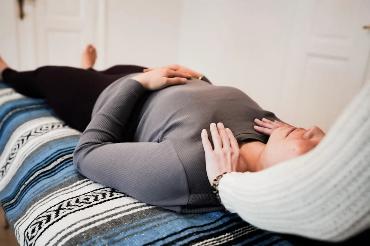 A person lying on a bed with another person gently touching their abdomen and head.