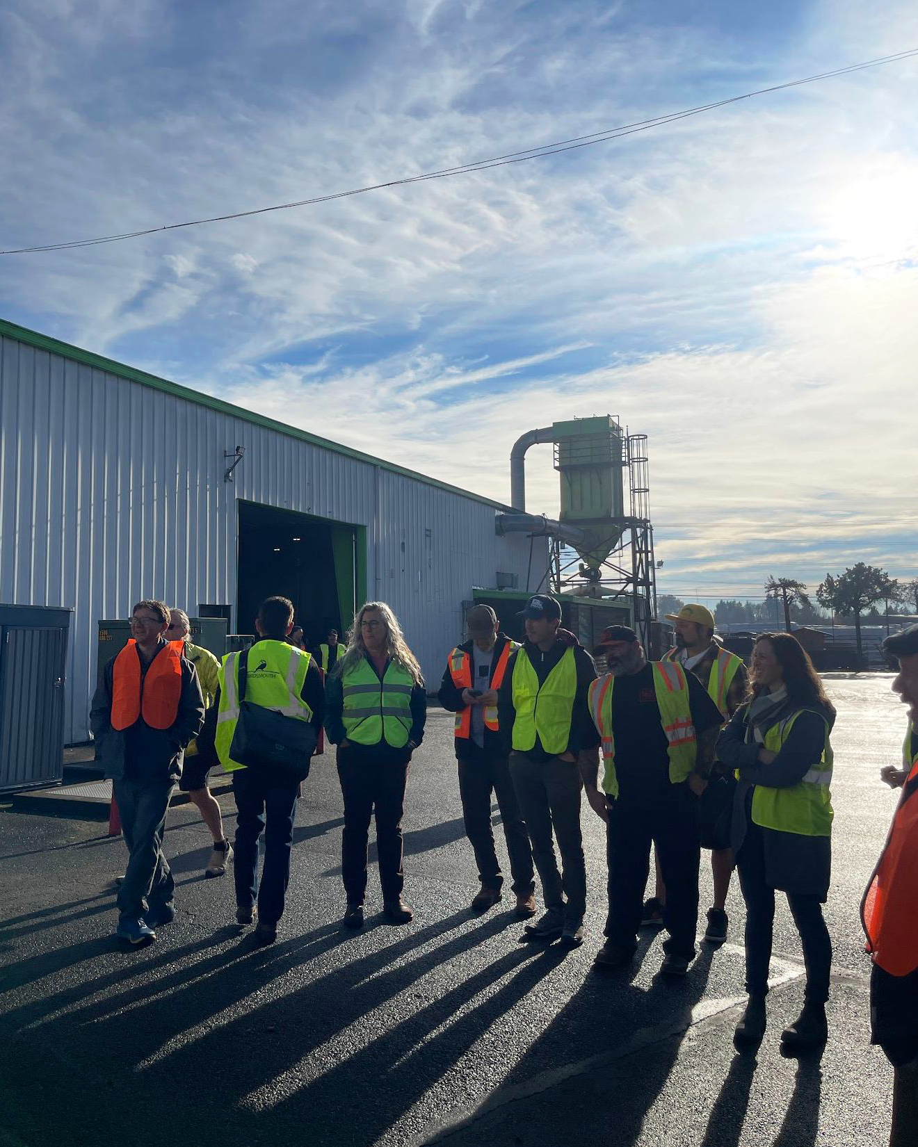 A group of people wearing safety vests and hard hats gathered outside a large industrial building.