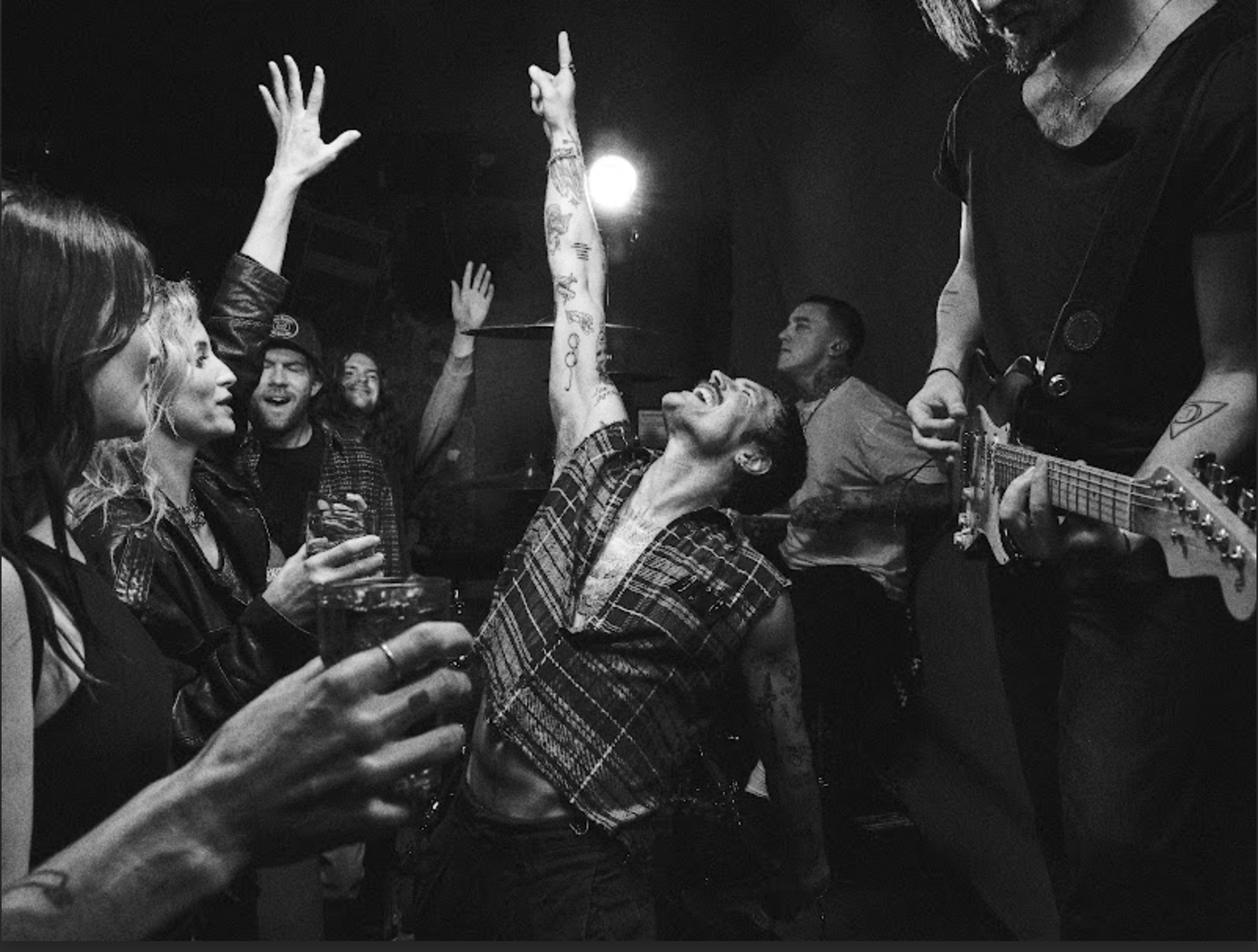 A lively black and white photo of a young woman dancing passionately at a concert or party, with people around her holding drinks and raising their hands, and a man playing guitar beside her.
