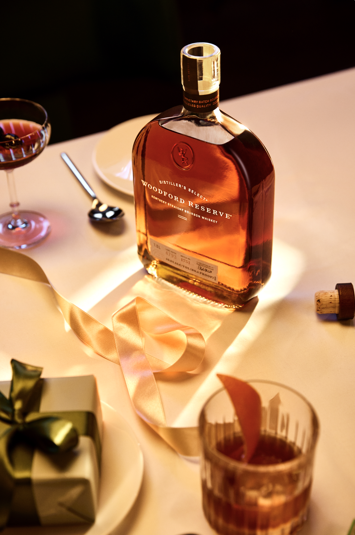 A bottle of Woodford Reserve bourbon whiskey on a table with a cocktail glass, a drinking glass with a slice of orange, a small gift box with a green bow, a pink ribbon, a plate, and a cork.
