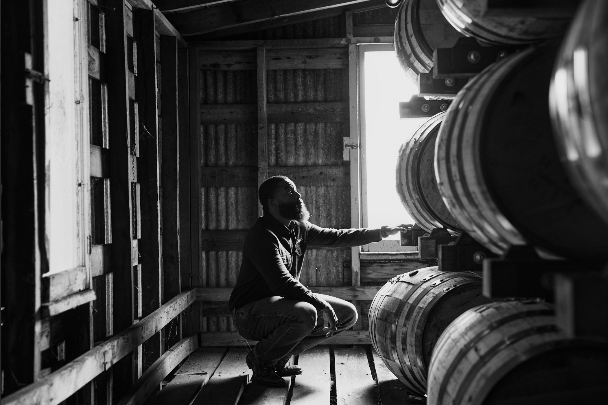 A man crouching inside a wooden storage room, reaching out towards barrels, illuminated by sunlight coming through a window.