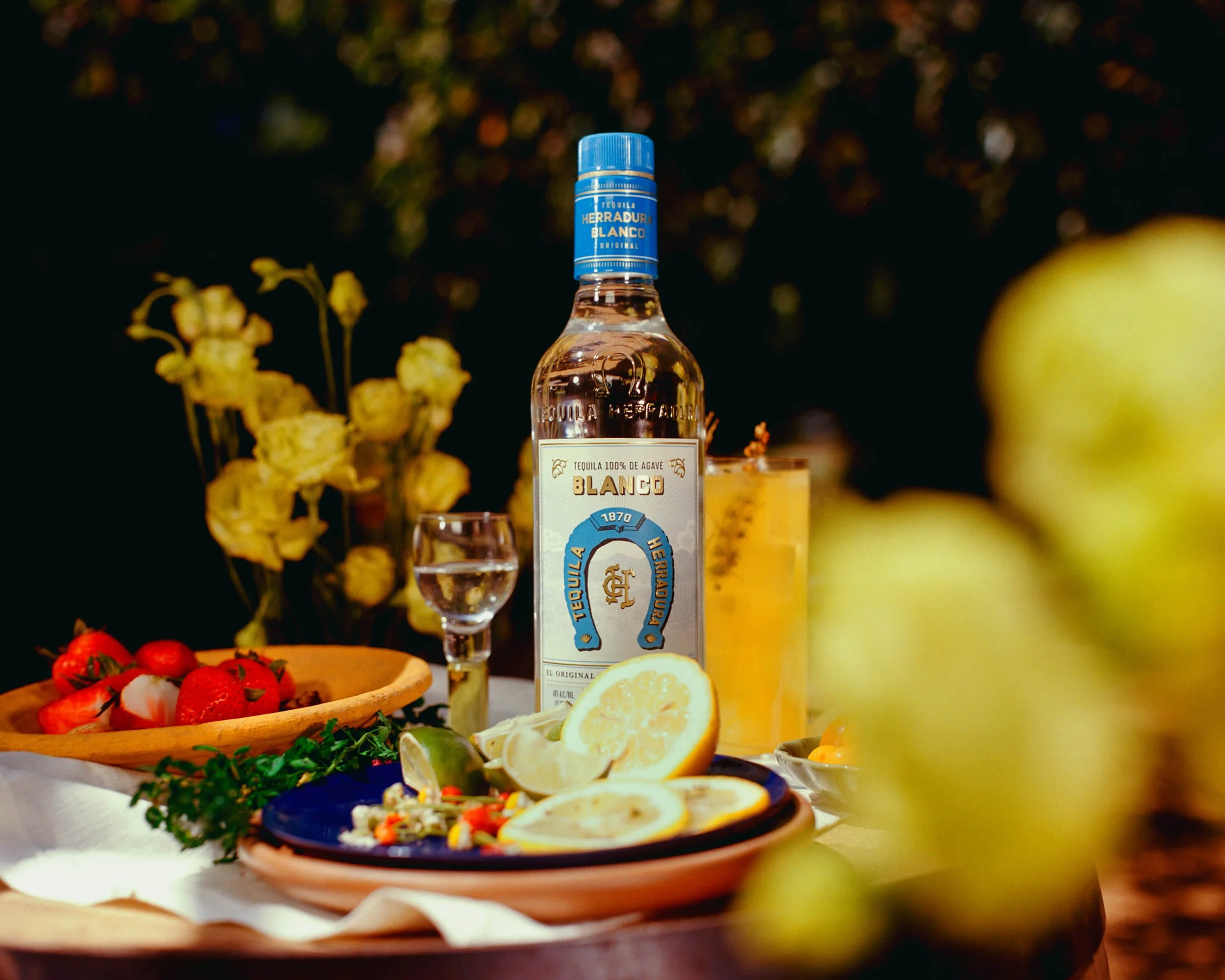 A bottle of Herradura Blanco tequila on a table with lemon slices, strawberries, and a small glass of tequila, surrounded by flowers and fruits outdoors.