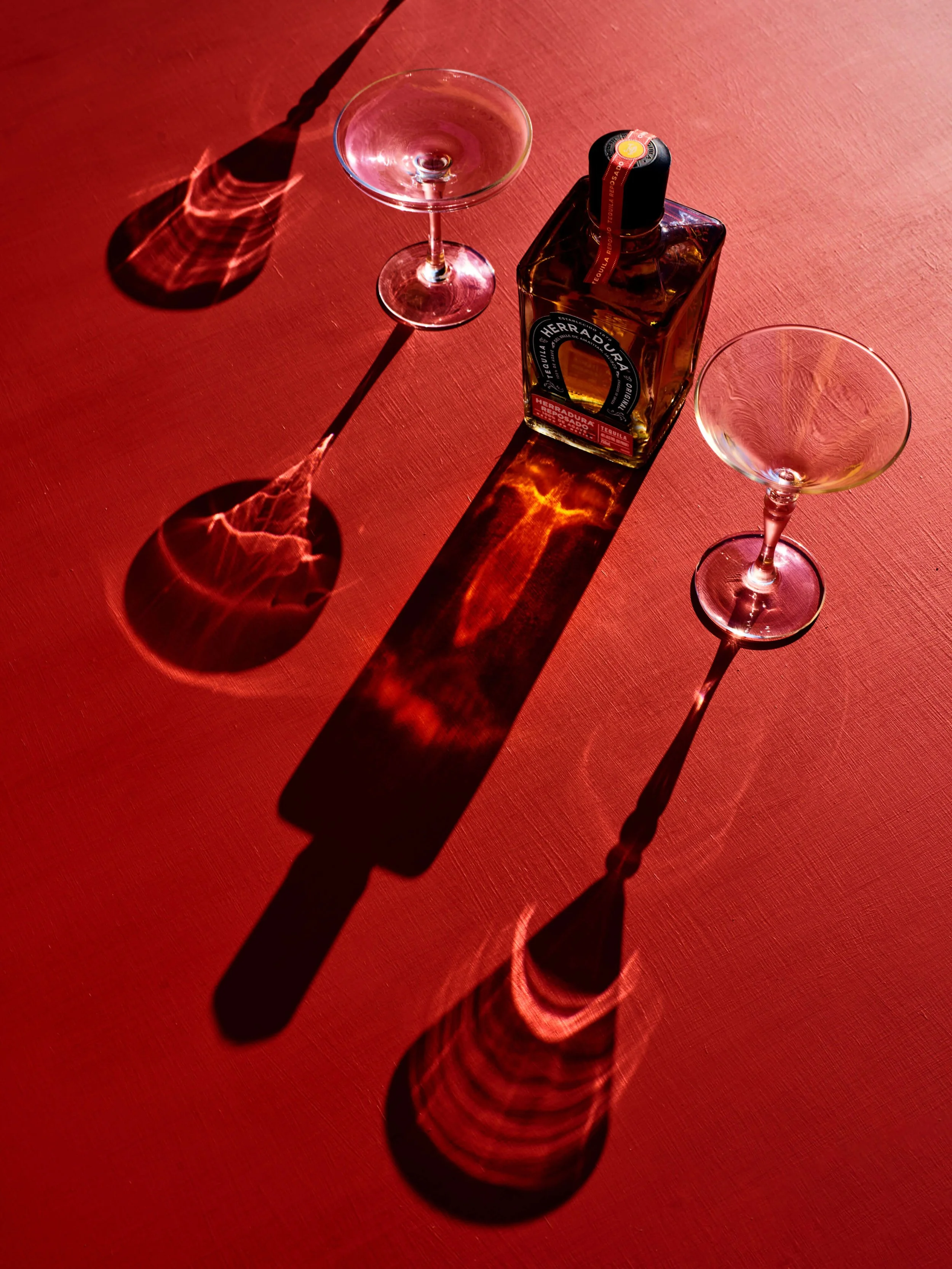 A bottle of Herradura tequila with two empty cocktail glasses on a red surface, casting colorful shadows from overhead lighting.