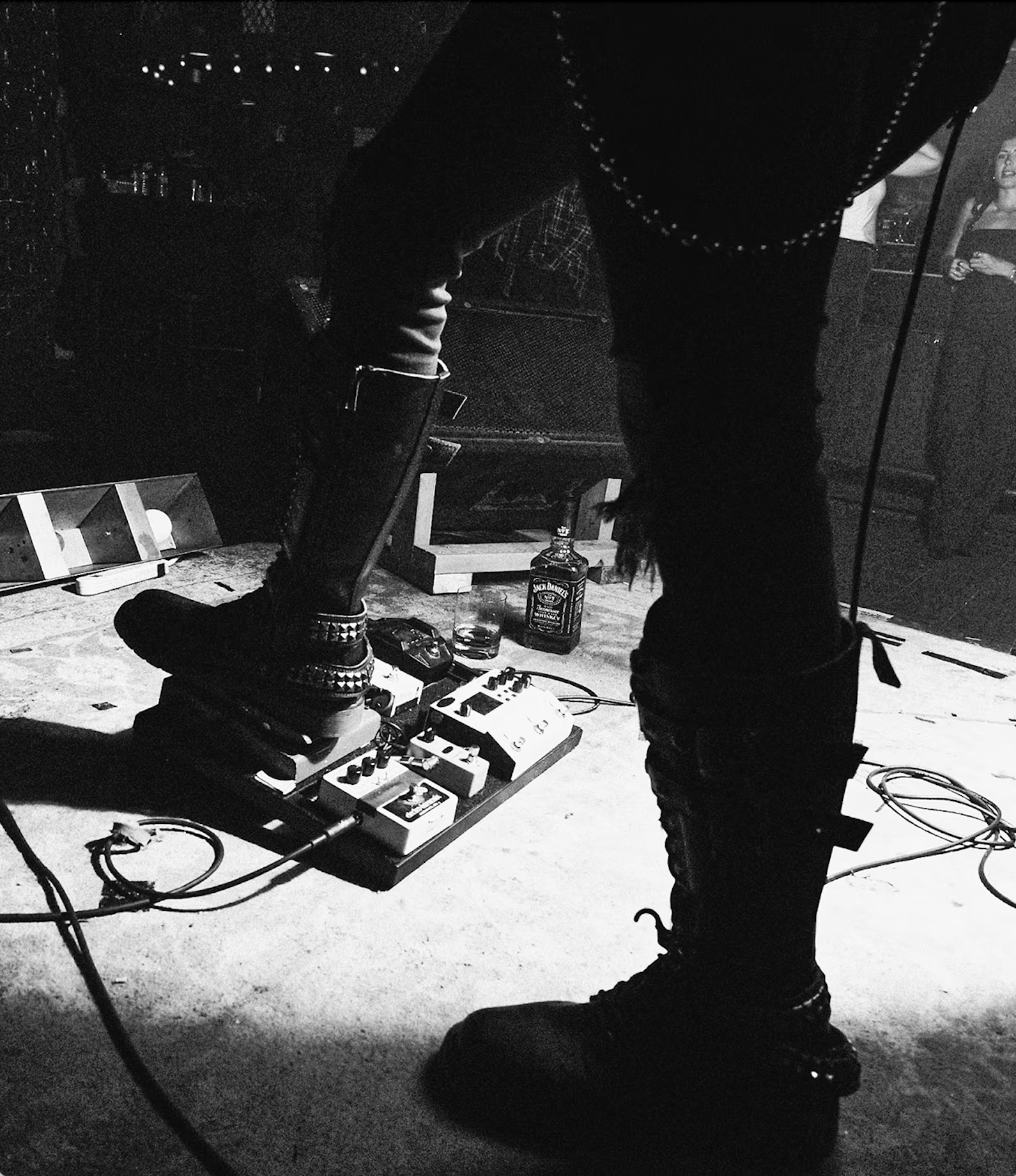 Black and white photo of a person standing on stage with boots and torn jeans, adjusting effects pedals on the floor, with a person in the background holding a drink in a dimly lit venue.