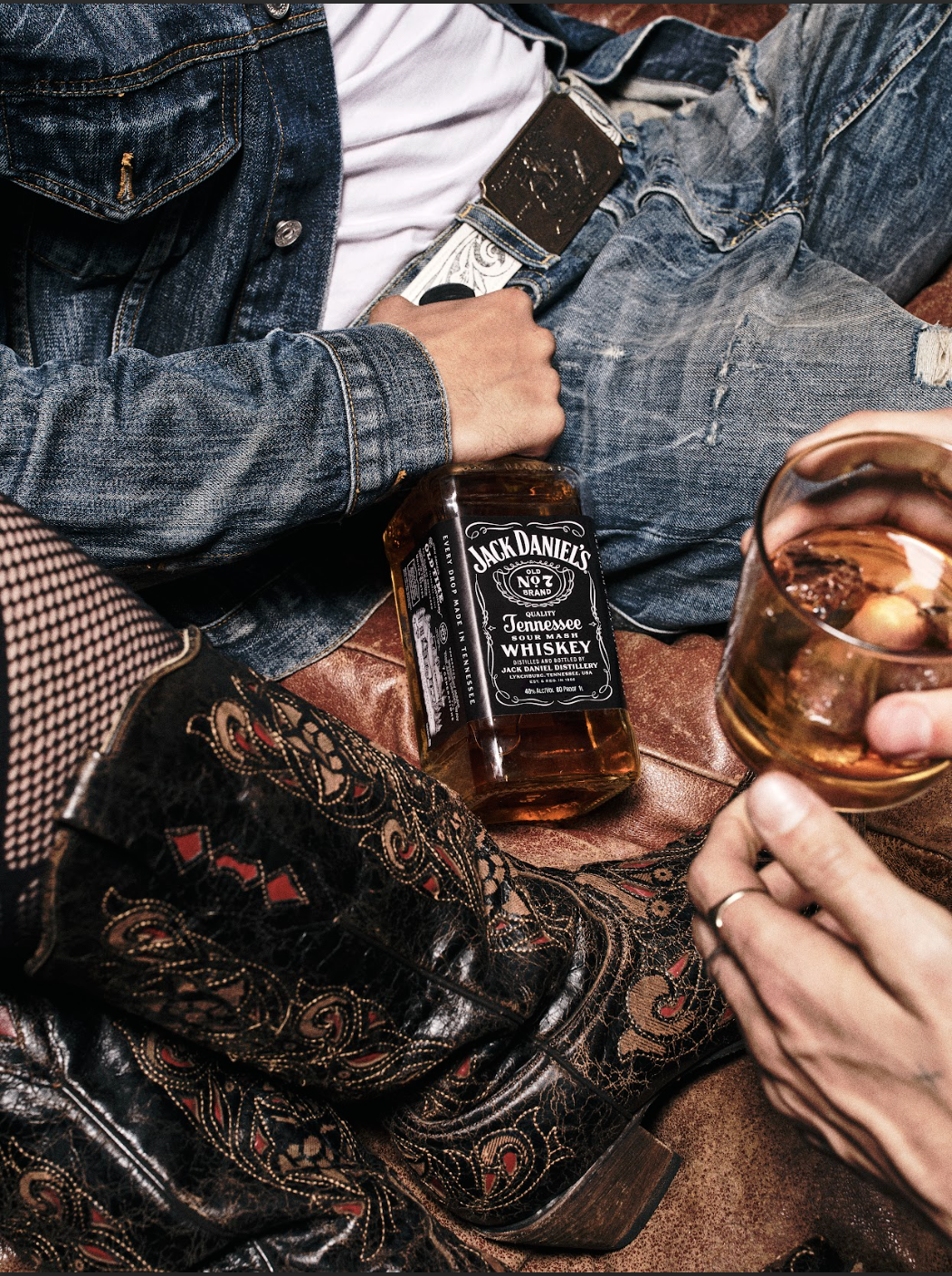 People sitting together with one person holding a glass of whiskey and a bottle of Jack Daniel's Tennessee whiskey resting on the floor, wearing denim jeans and a leather jacket.