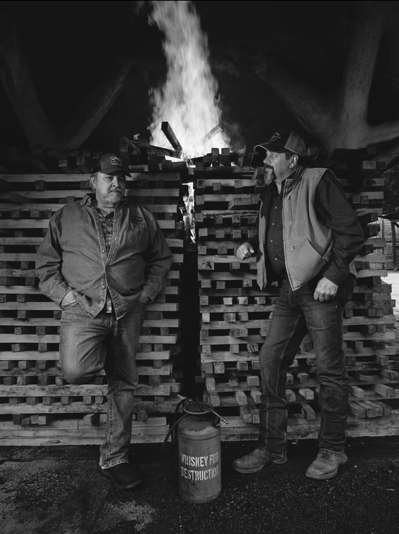 Two men standing in front of a woodpile with a fire burning behind, one with hands in pockets and the other talking, a fire extinguisher labeled 'Whiskey for Destruction' on the ground.