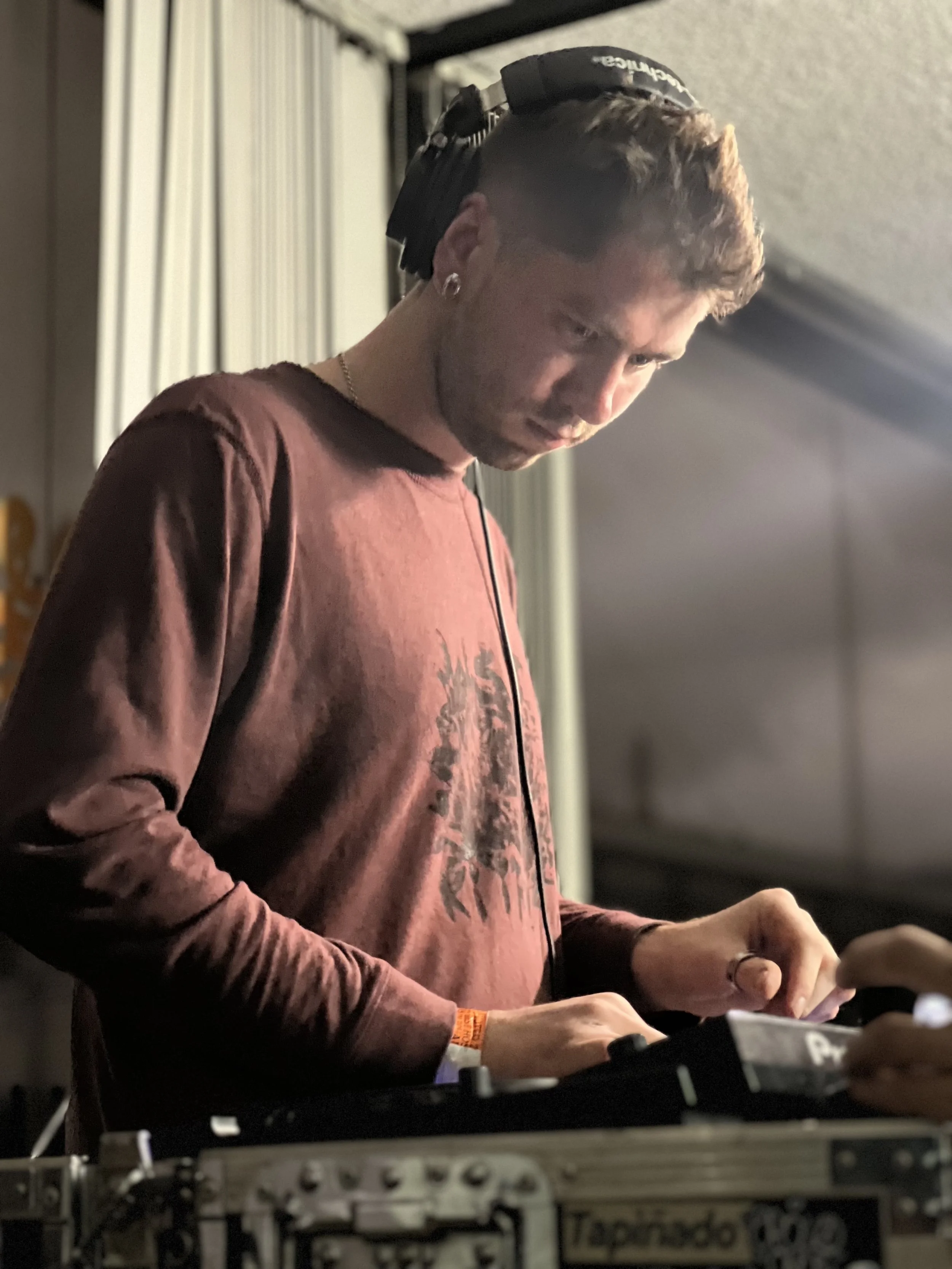A male DJ wearing headphones and a maroon long sleeve shirt, focused on mixing music at a DJ controller in a dimly lit indoor setting.