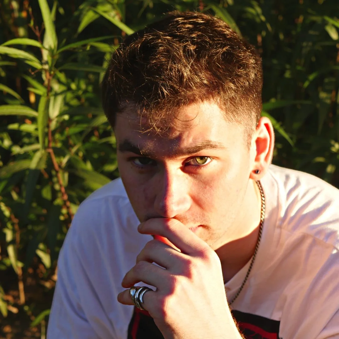 A young man with short brown hair and piercing green eyes, wearing a white shirt, jewelry, and rings, looking at the camera with a serious expression in a lush outdoor setting with green plants and sunlight.