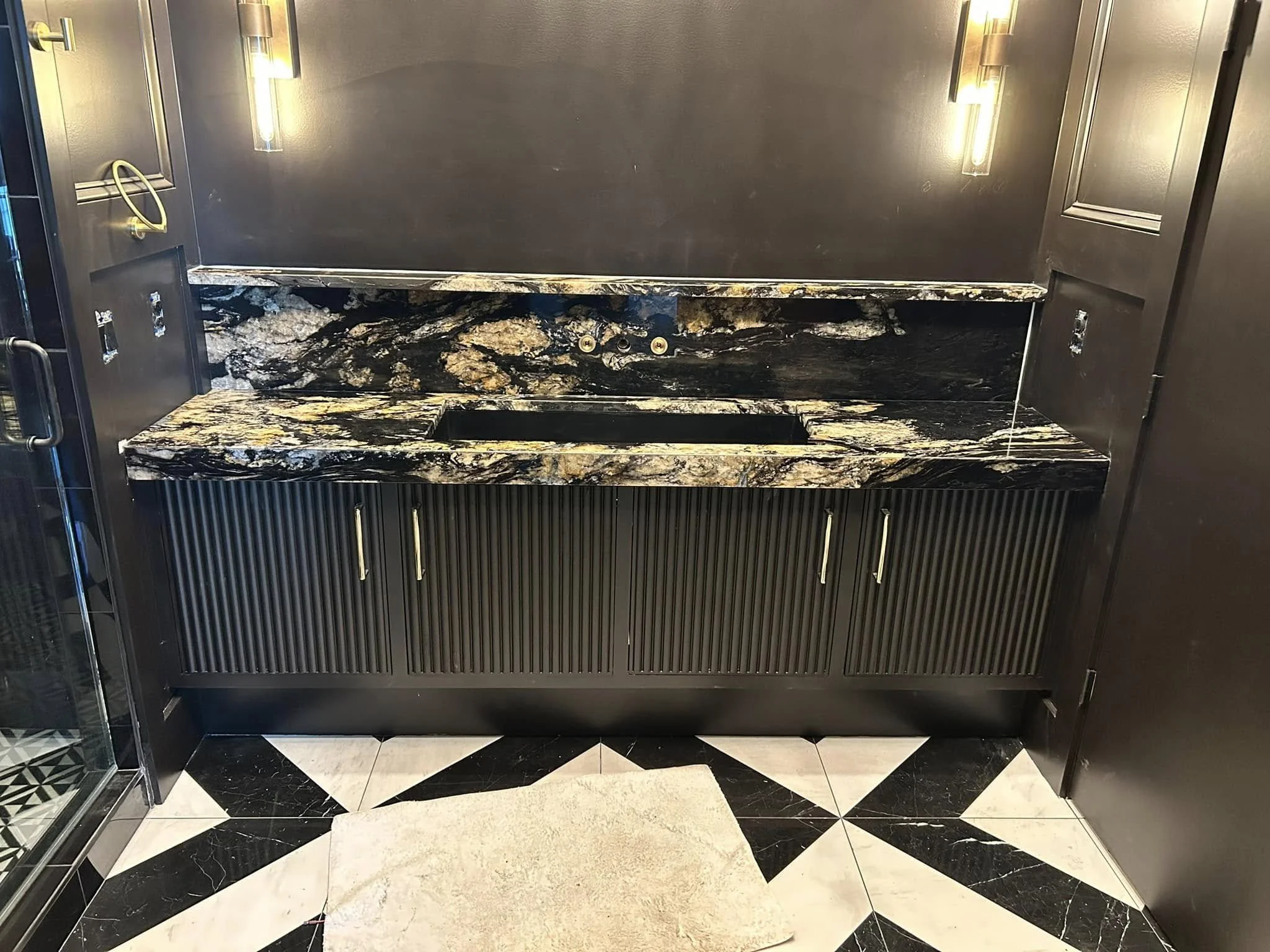 Black and gold marble countertop with built-in space for a sink, flanked by black cabinets with vertical grooves, and illuminated by two wall-mounted lights in a modern kitchen or bar area.