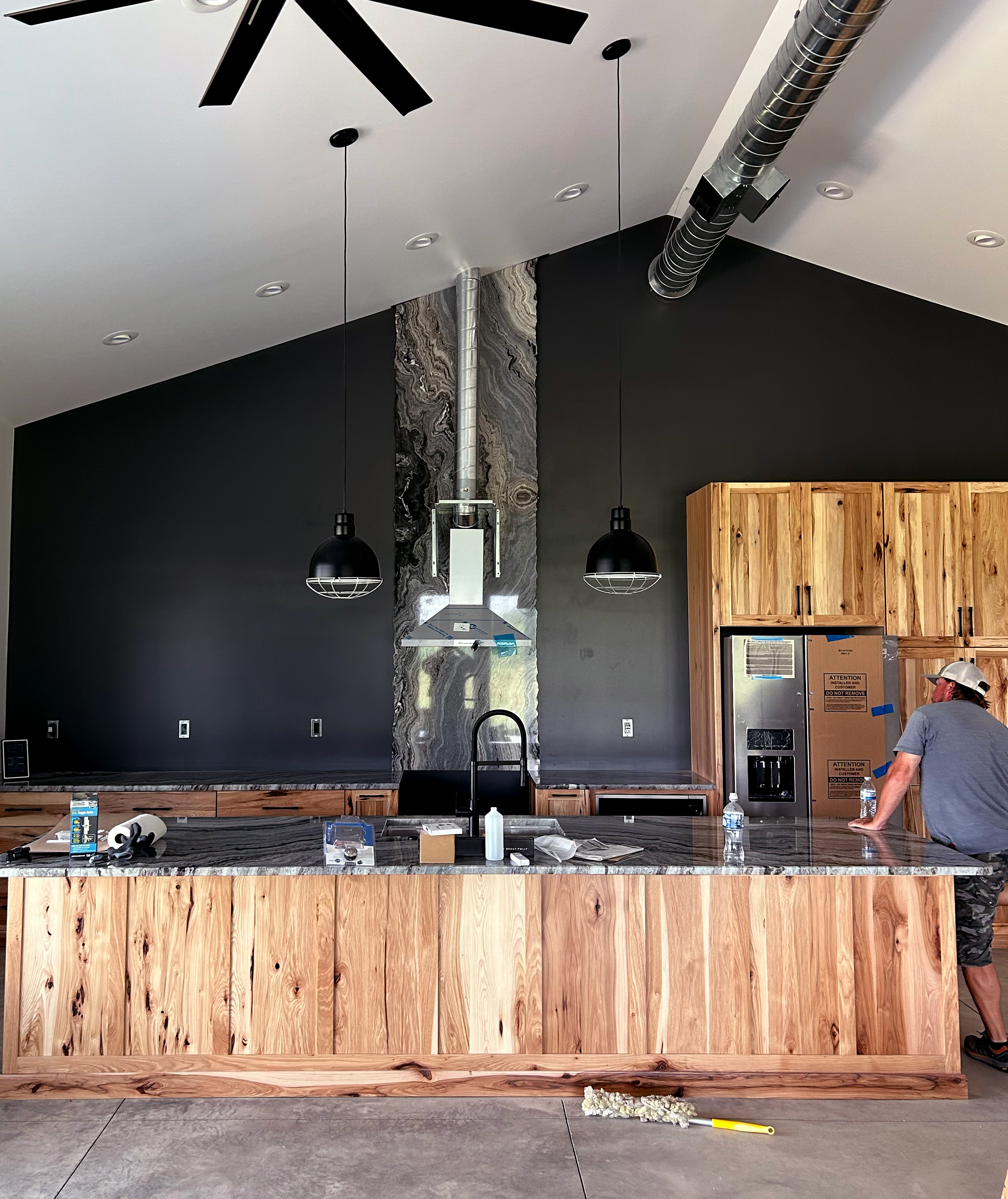 Modern kitchen with wooden cabinets, black backsplash, marble countertop, and two black pendant lights. A man stands at the counter with tools and water bottles nearby, indicating ongoing installation or renovation.