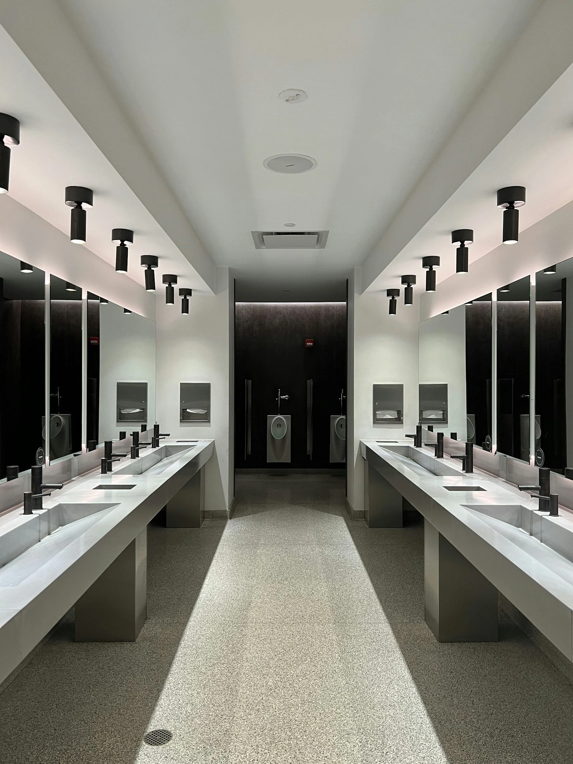 Empty public restroom with double sinks, mirrors, soap dispensers, and hand dryers, well-lit with modern lighting fixtures.