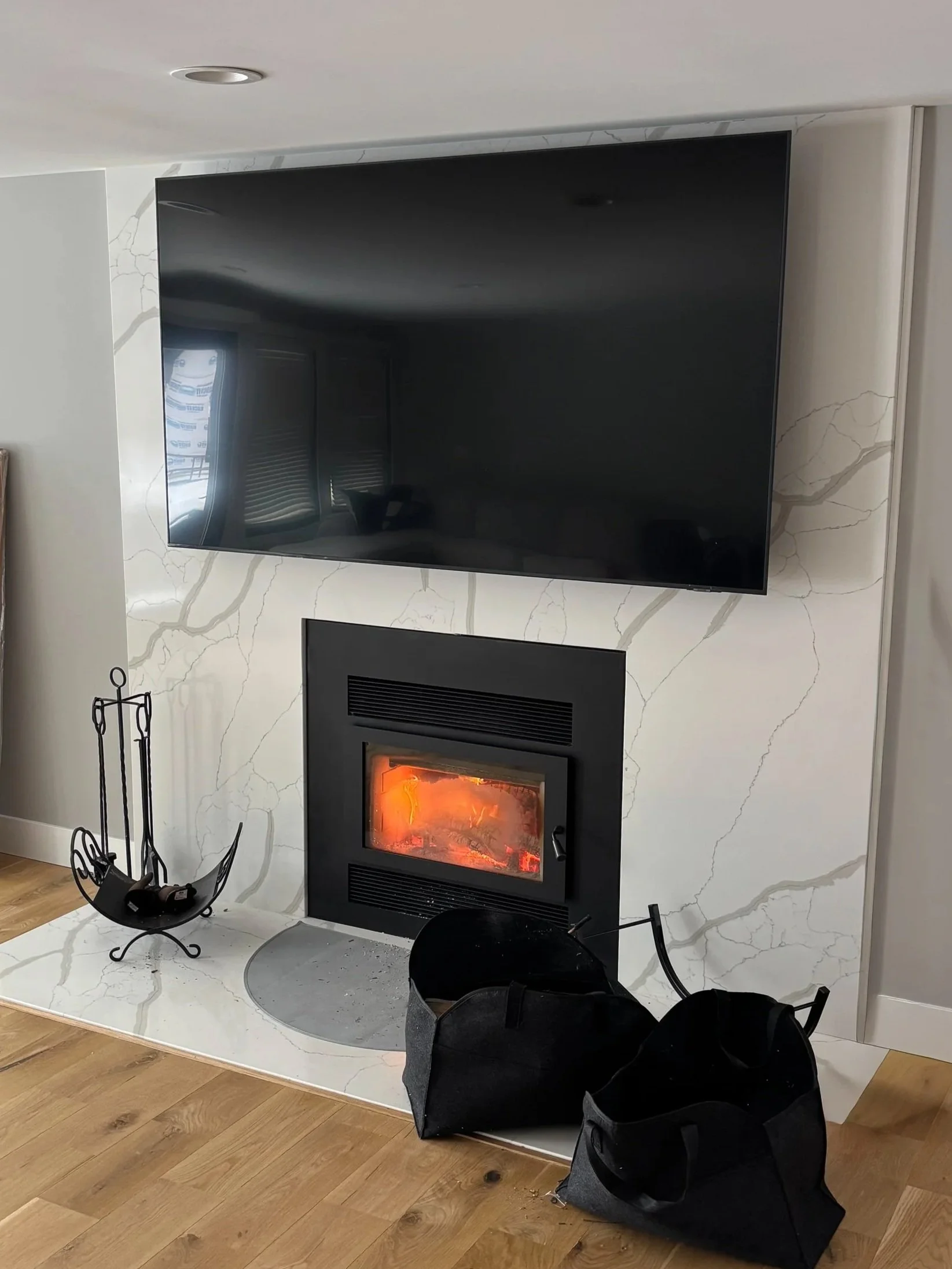 Modern living room corner with a flat-screen TV mounted on a marble wall above a fireplace, with a black fire surround and tools, and two black bags on the hardwood floor.