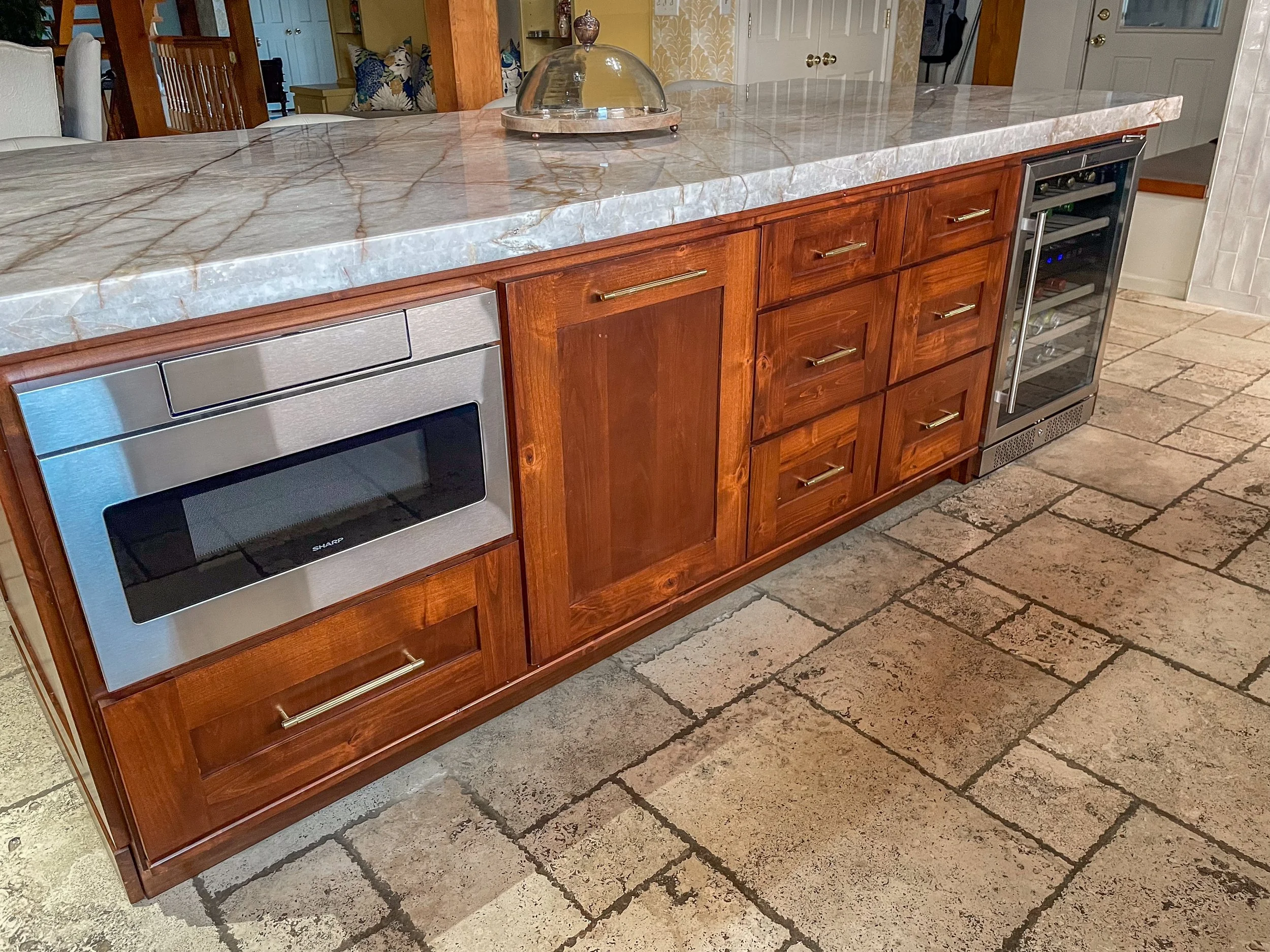 Kitchen island with a marble countertop, built-in microwave, and wine cooler, with wooden cabinets and large stone tiles on the floor.