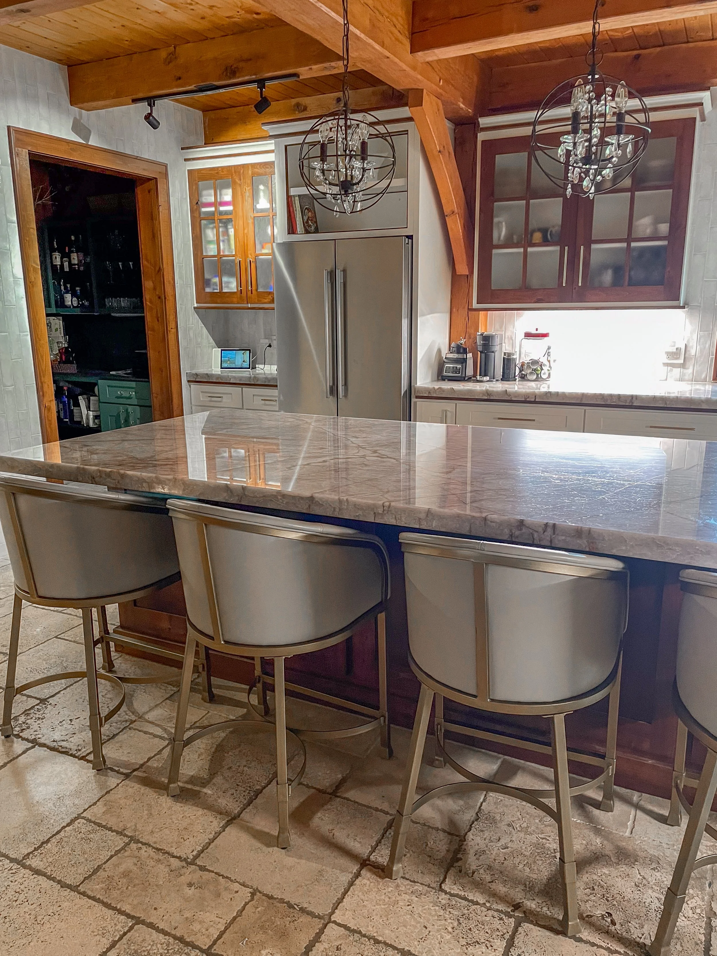 A kitchen with a marble island, three chairs, a stainless steel refrigerator, wooden cabinets, whitish walls, a tiled floor, and two chandeliers hanging from a wooden ceiling.